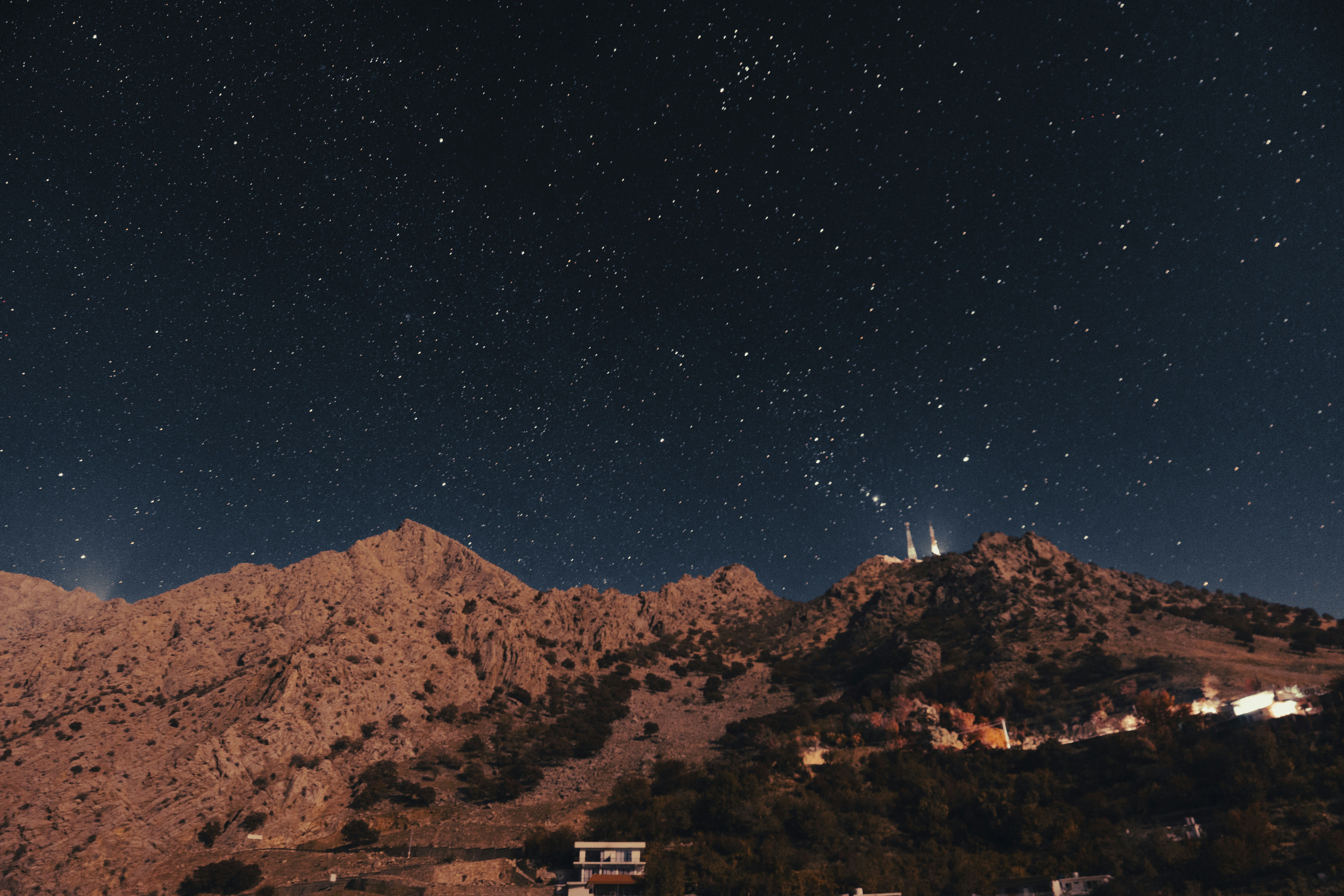 Starry night sky over rugged mountains with lights.