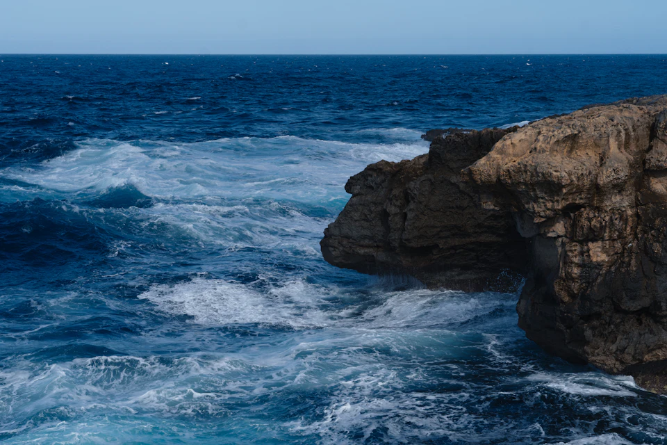 The image depicts a rocky cliff face with a deep blue ocean stretching out to its right side, where waves are crashing against the rocks and creating white foam on the surface. The sky above is light blue, suggesting a clear day. In the distance, there are several boats visible in the water, adding depth to the scene.