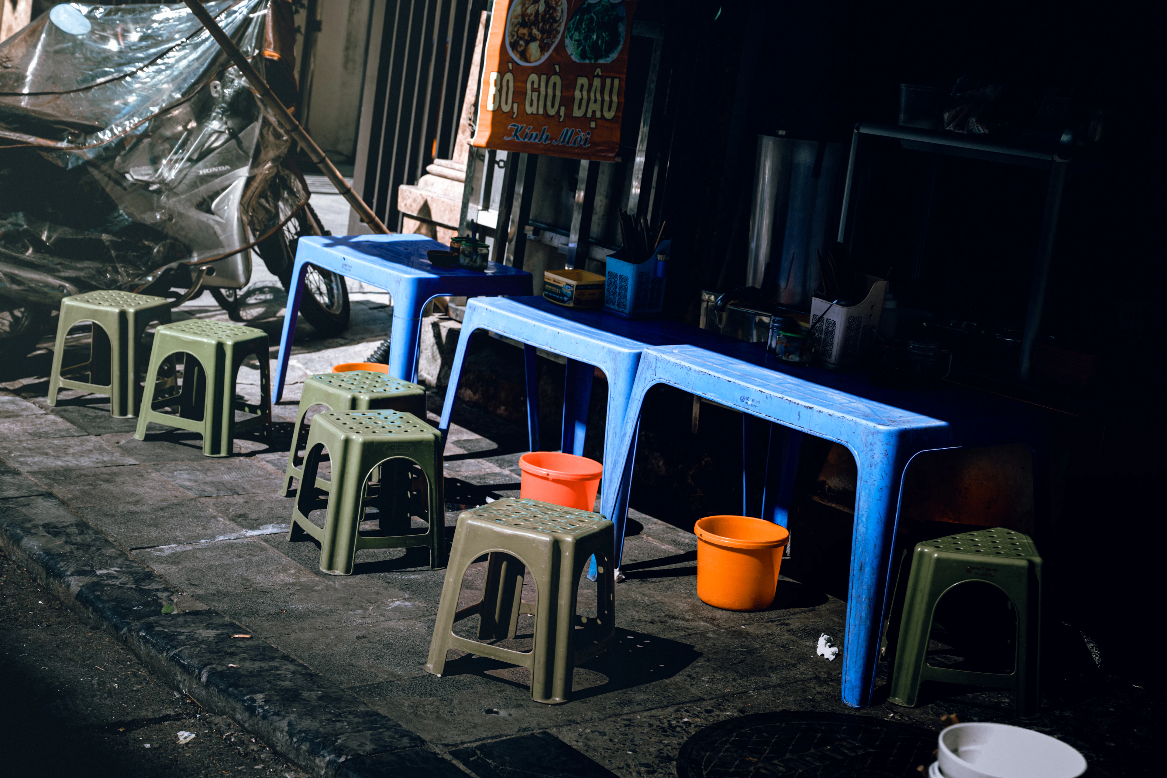 Low blue plastic tables and small stools set out on the pavement outside a dark Hanoi street-food shopfront, orange buckets tucked between them and a hand-lettered Vietnamese menu board on the wall above.