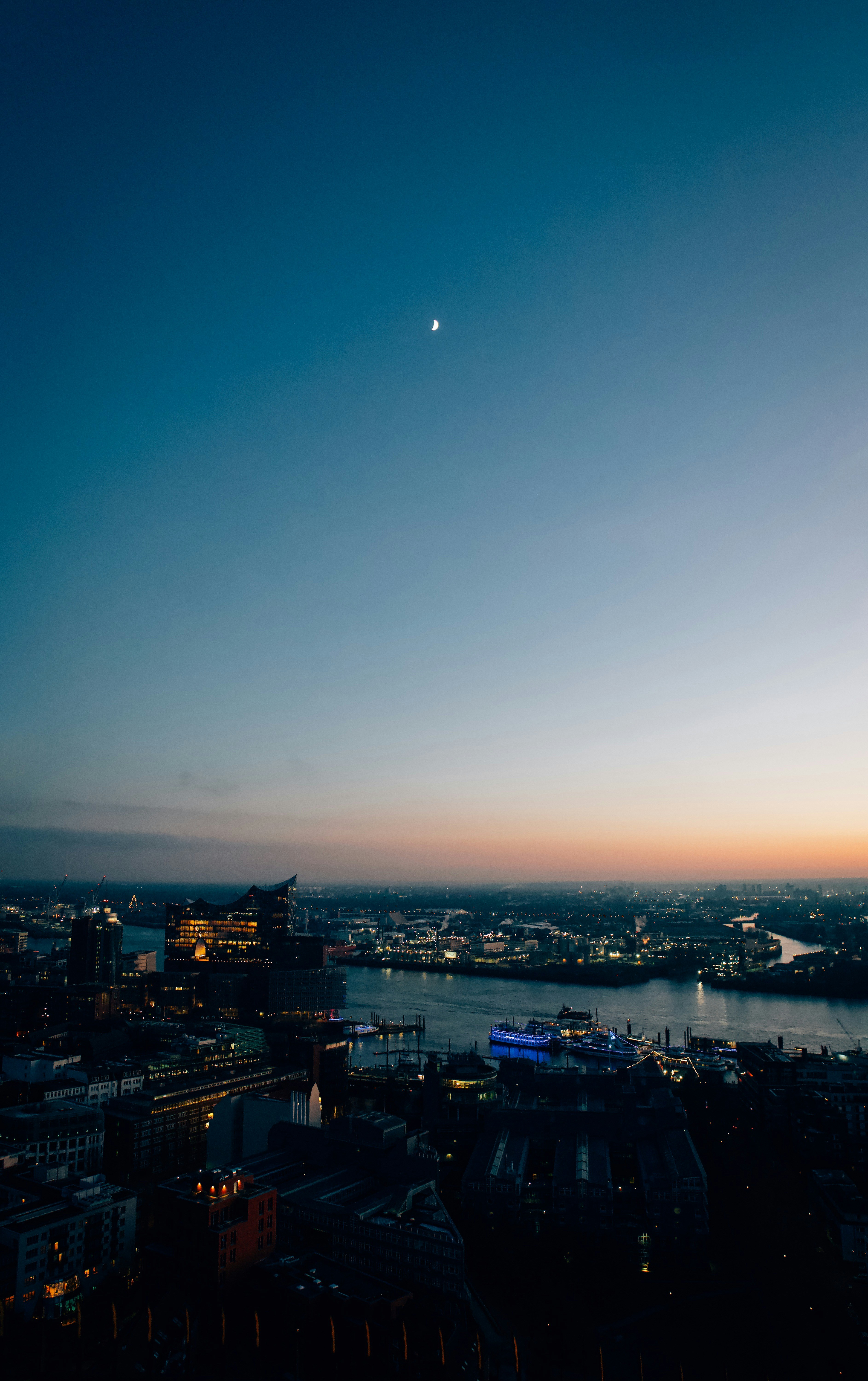 Cityscape at dusk with moon and harbor lights.