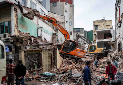 Excavators demolish buildings amidst rubble and onlookers.