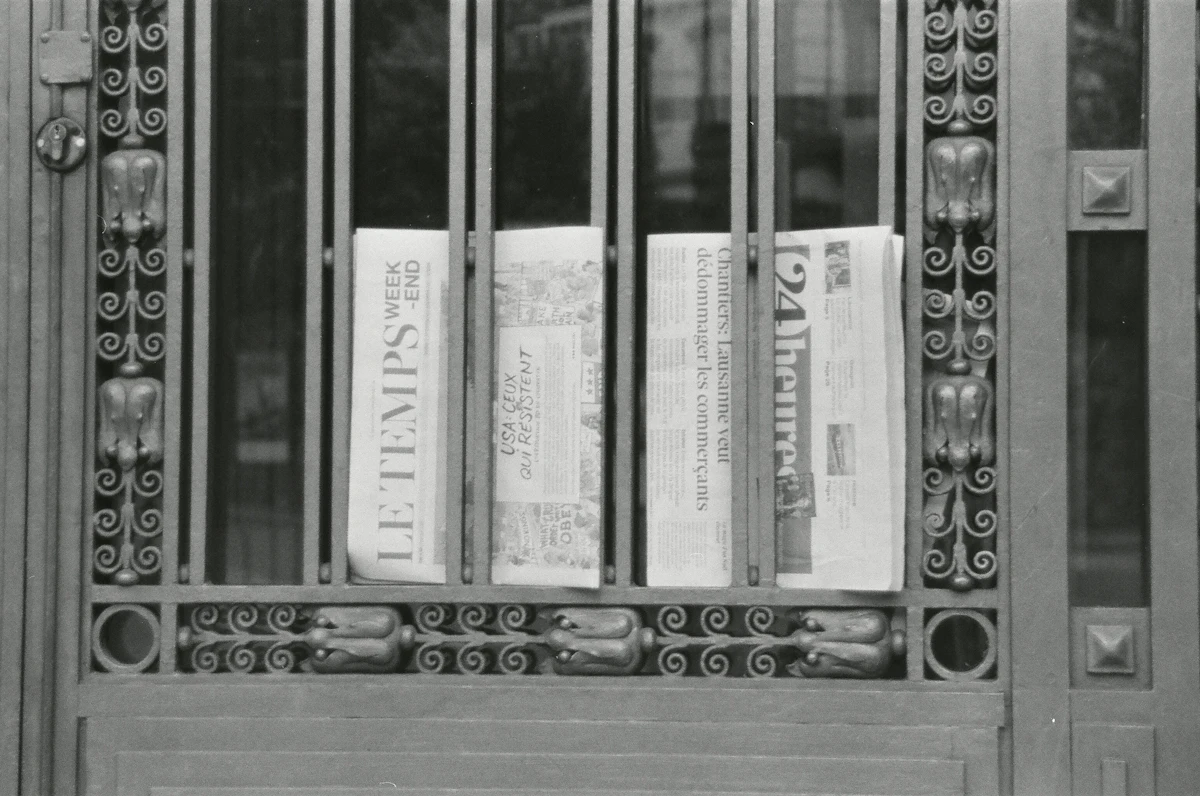 Newspapers stacked behind an ornate metal gate — a symbol of locked-away local information