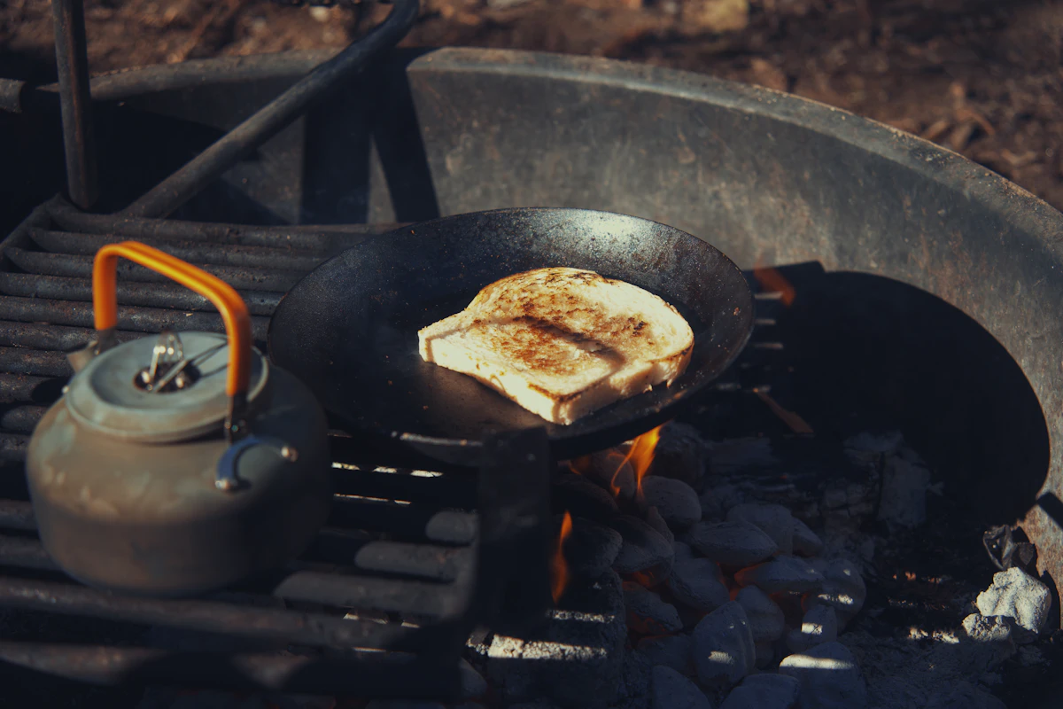 Bread browning in a steel pan over campfire coals, kettle warming alongside