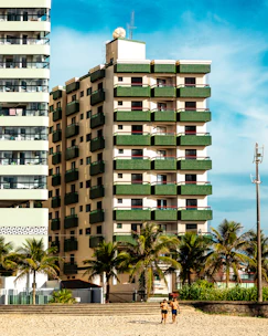 Two people walk on a sandy beach near buildings.