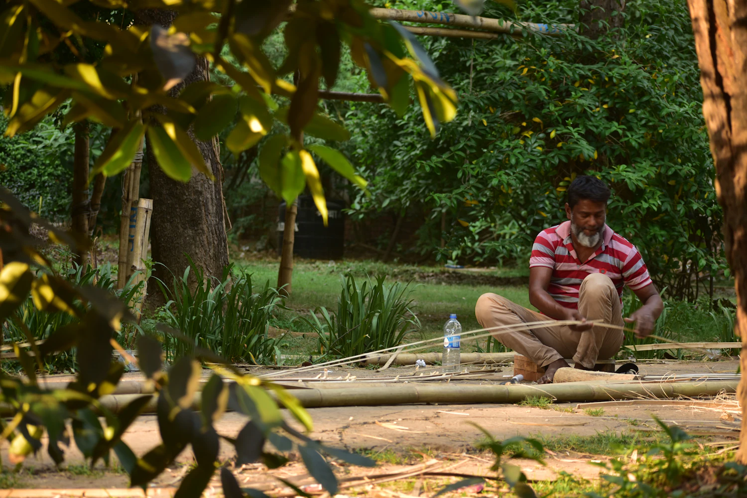 A man sitting outdoors in the shade working a length of bamboo