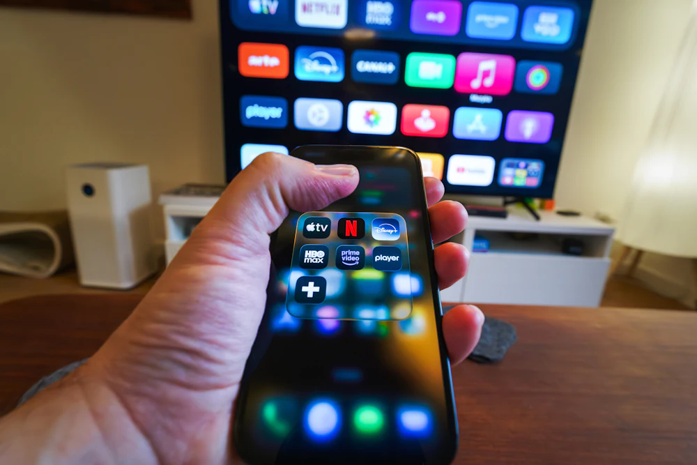Hand holding a smartphone with streaming service apps in front of a television