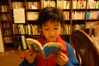 A young child reads a book in front of bookshelves.