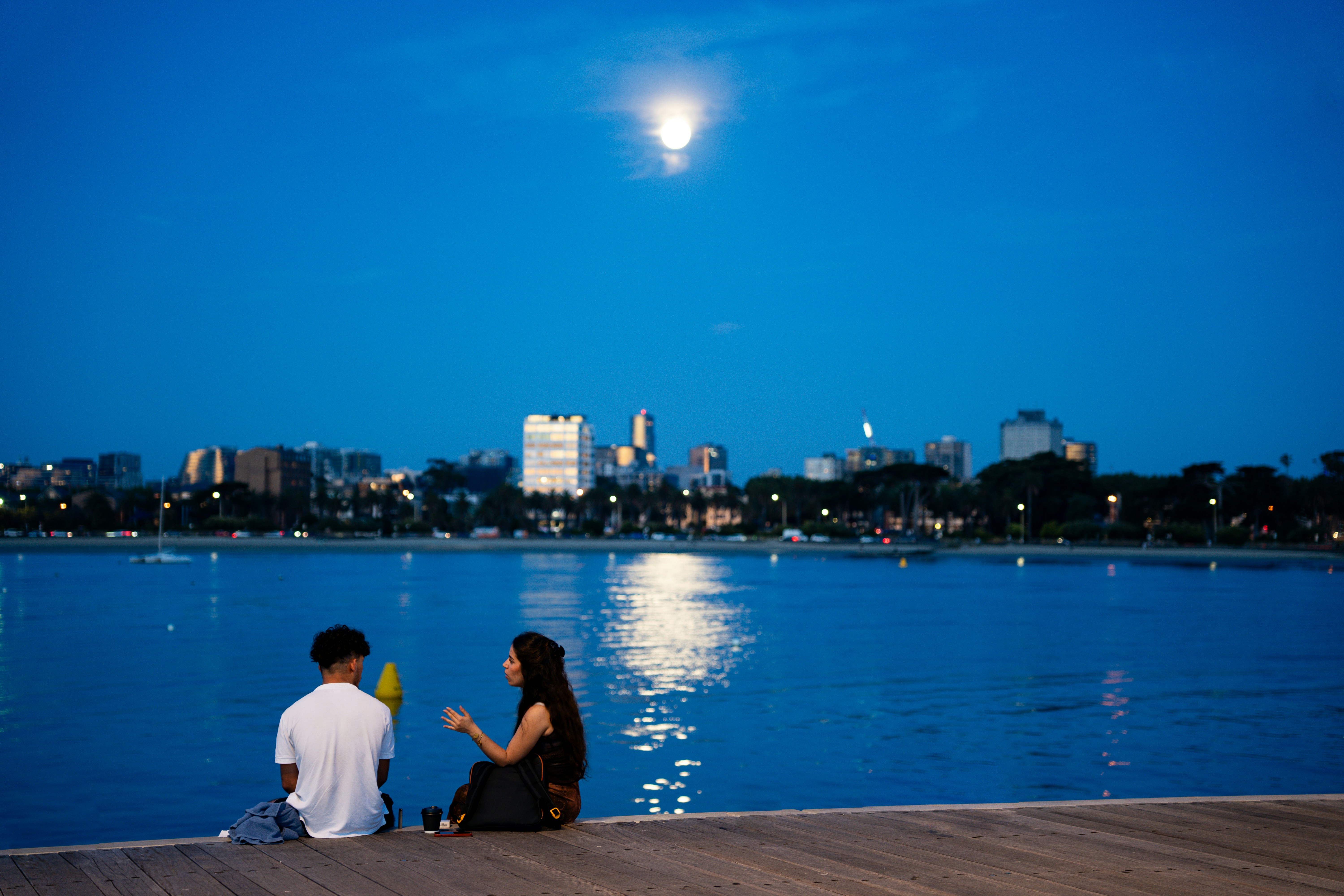Couple sitting by water with city skyline at night.