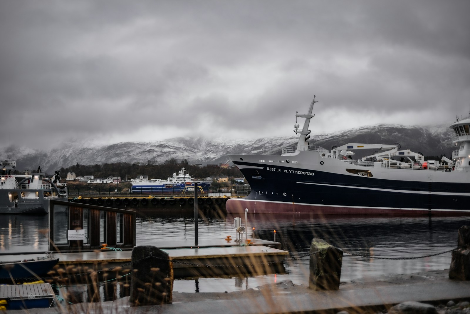 A fishing vessel moored in a Norwegian harbour under heavy cloud, with a snow-covered ridge behind it