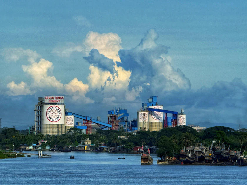 An industrial complex on a riverbank under a cloudy sky in Chattogram, Bangladesh.