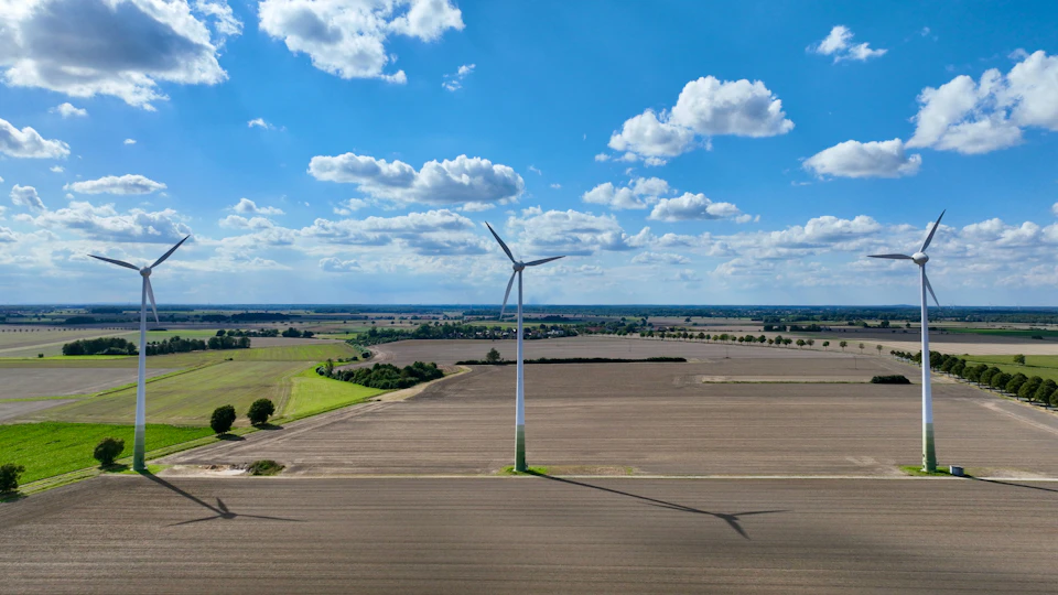 Three towering white wind turbines stand against a clear blue sky with fluffy clouds