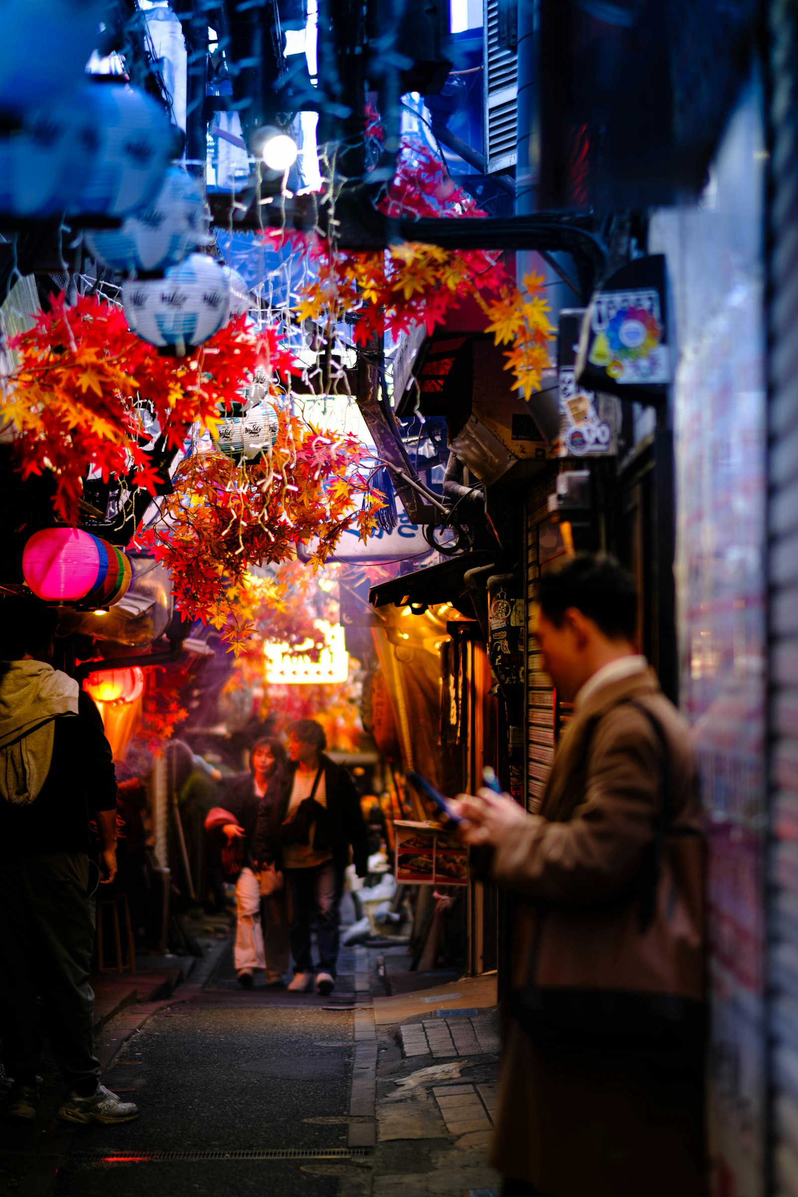 A man checking his phone in an izakaya alley decorated with lanterns and maple leaves