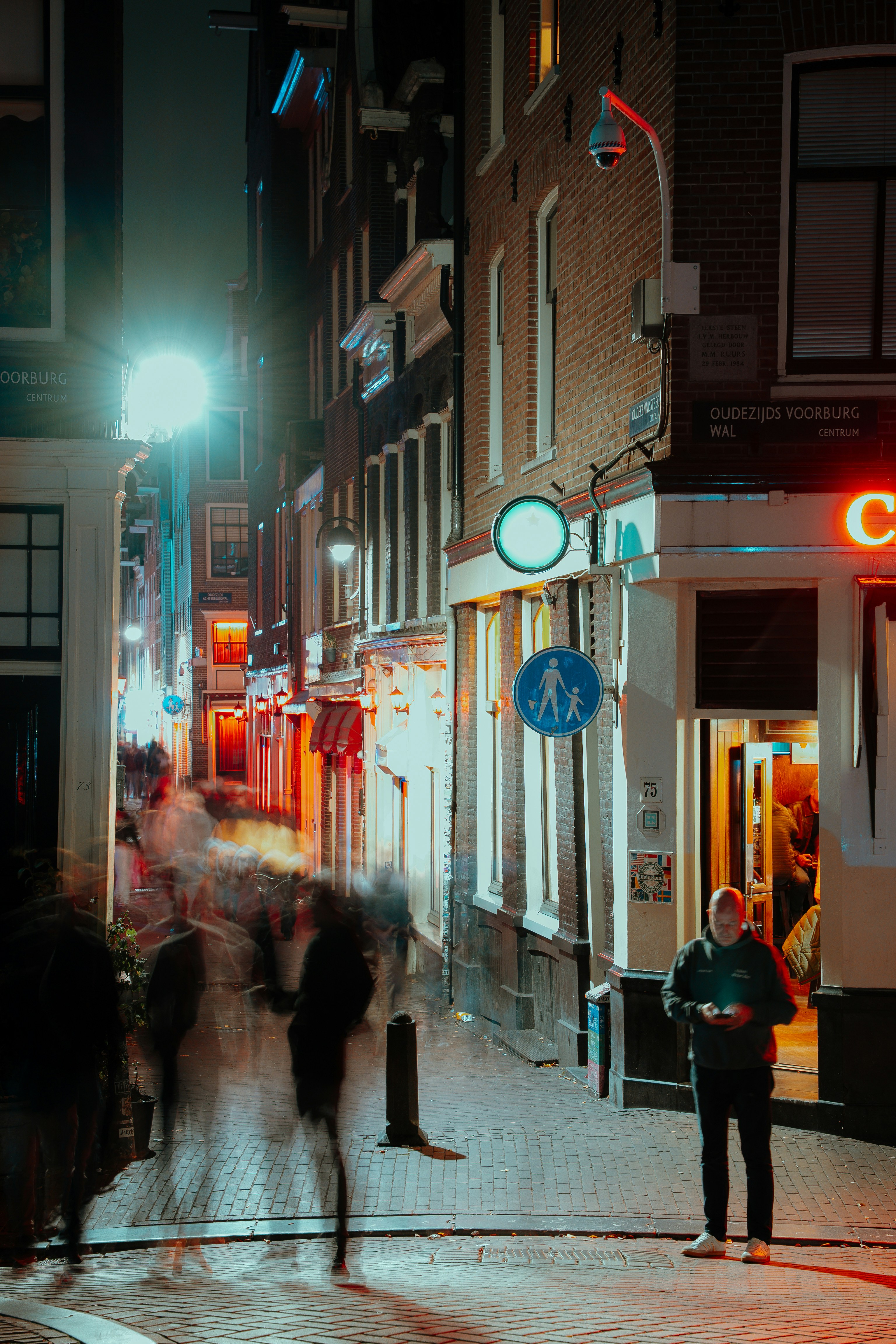 Figuras borradas caminham por uma rua estreita à noite.