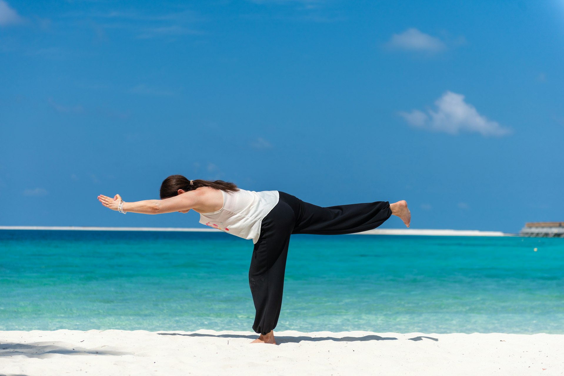 Woman balancing on one leg on a white sand beach in Warrior III, her arms and lifted leg stretched parallel to the ground