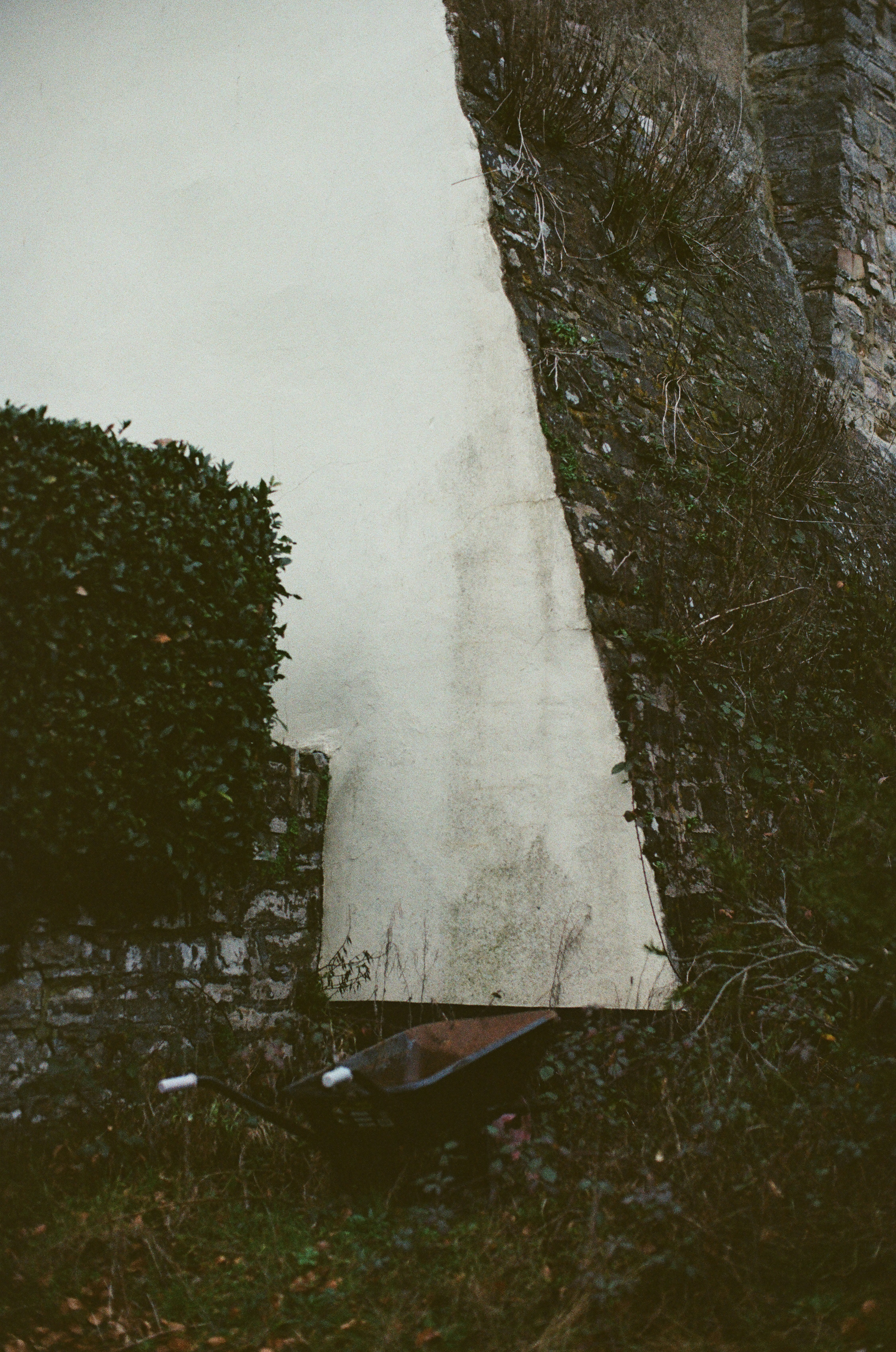 A rusty wheelbarrow rests near a stone wall.
