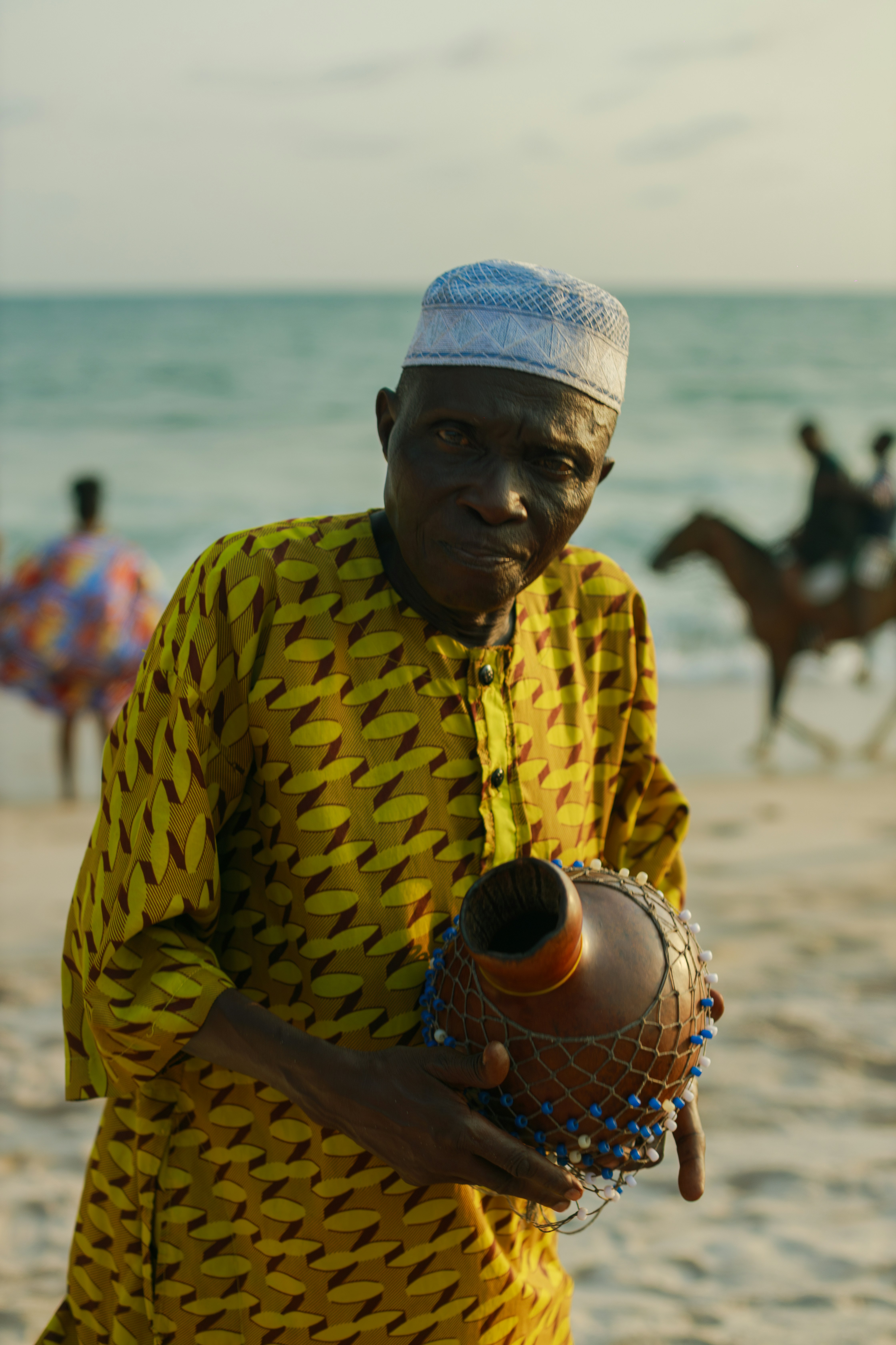 Man in traditional attire holding a musical instrument on beach.