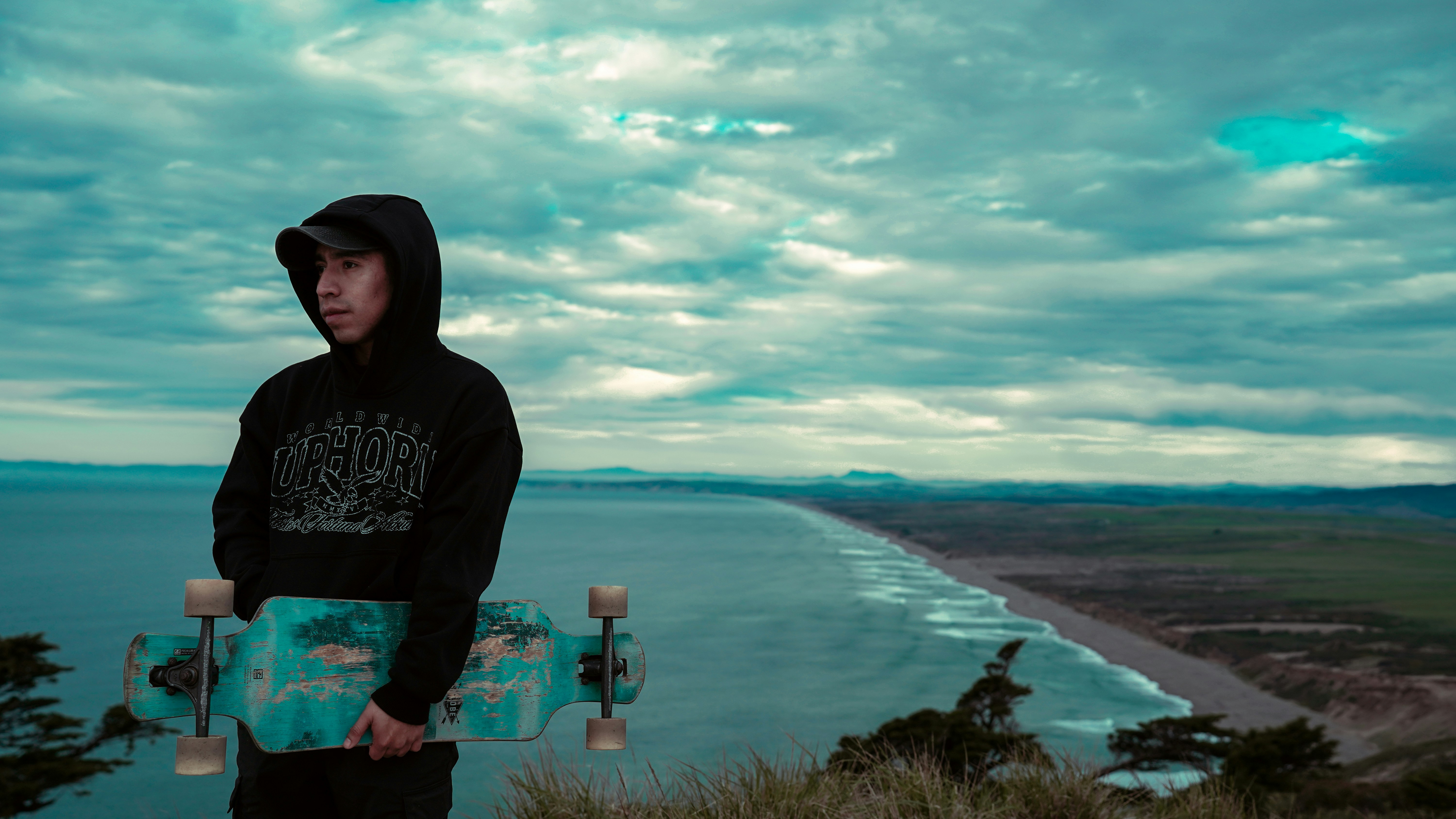 Junger Mann mit Skateboard auf einer Klippe mit Blick auf den Ozean