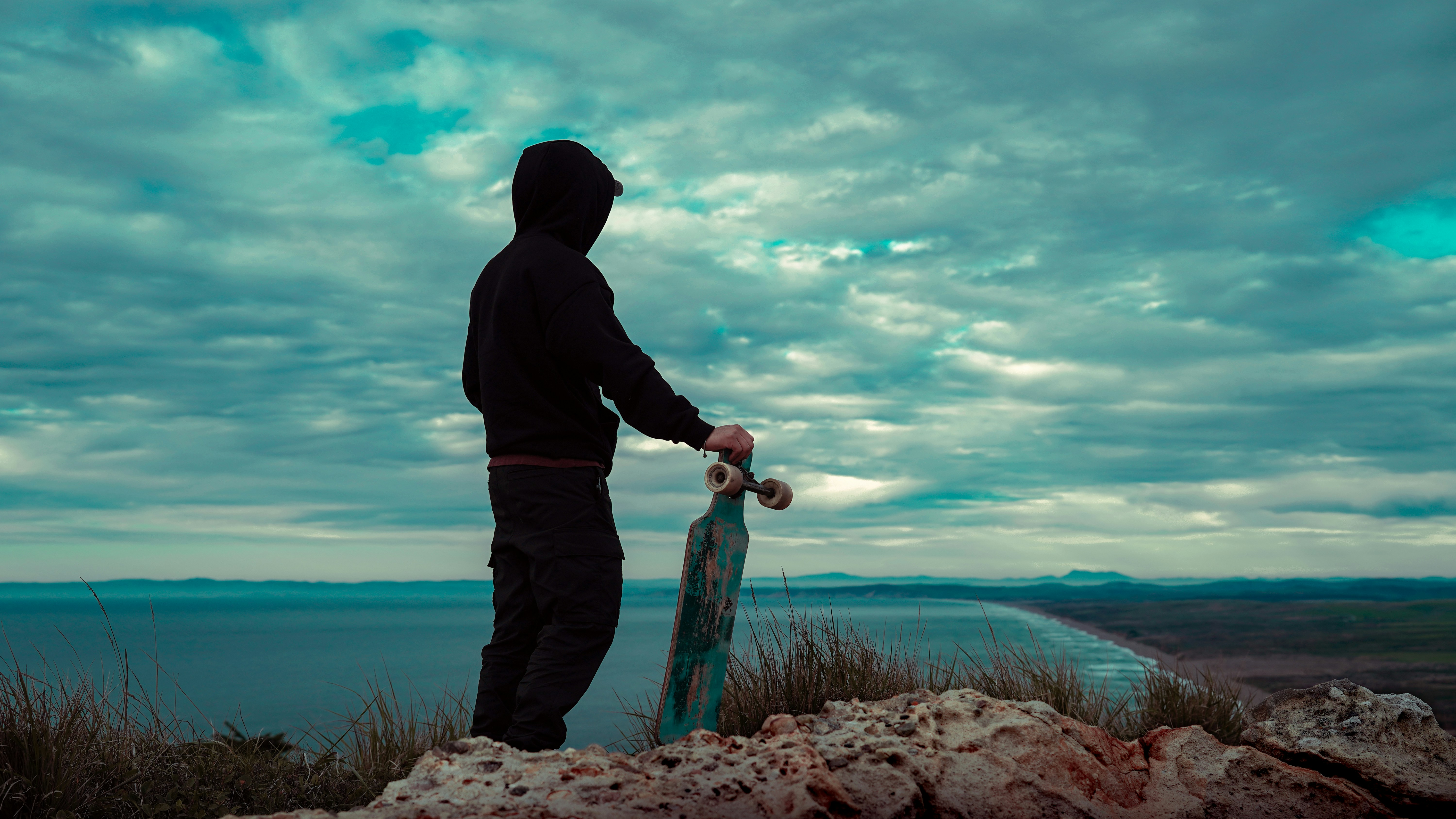 Person im Hoodie hält Skateboard auf einer Klippe mit Blick auf den Ozean