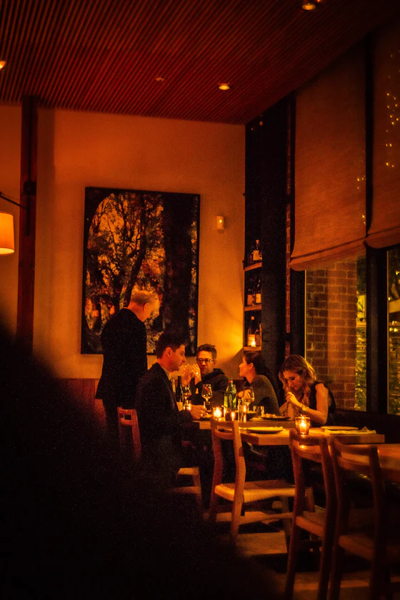 A warm amber dining room at night, candles on the tables, people eating and a waiter standing over them talking.