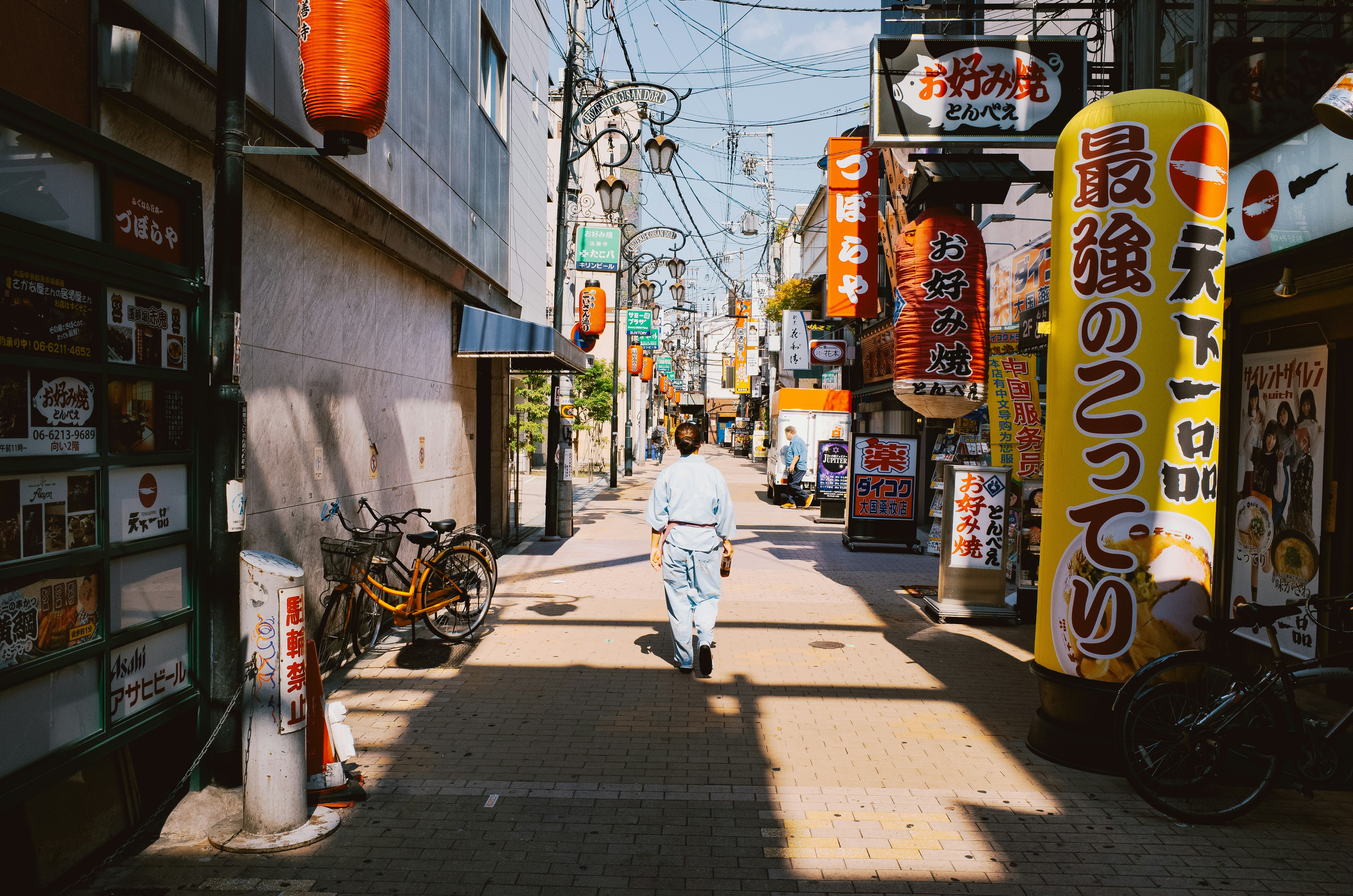 Person walks down a sunlit street with japanese signs.