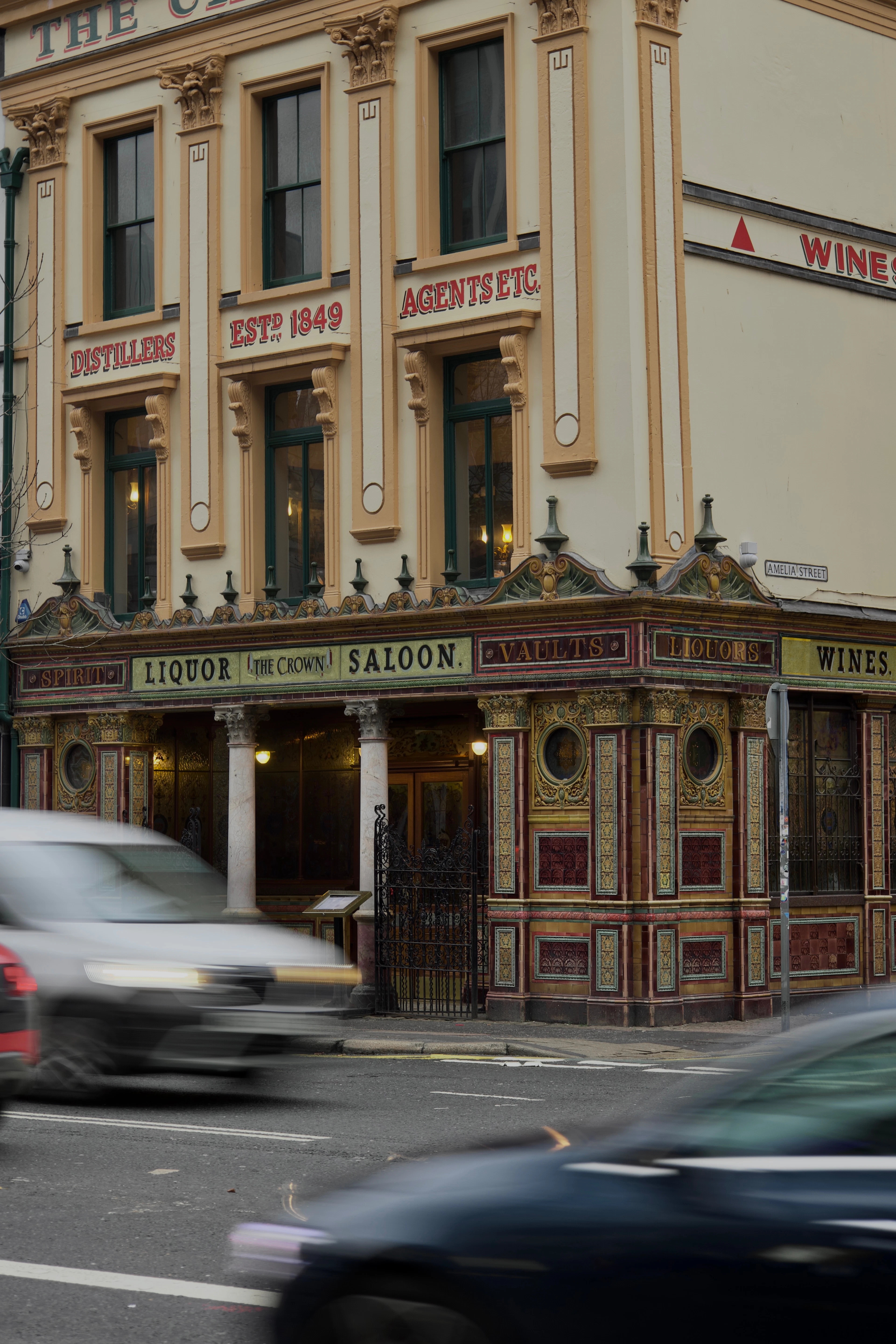 The tiled and gilded Victorian frontage of the Crown Liquor Saloon in Belfast, its painted lettering reading LIQUOR SALOON, VAULTS and WINES above ornate columns, with traffic blurring past on Great Victoria Street.