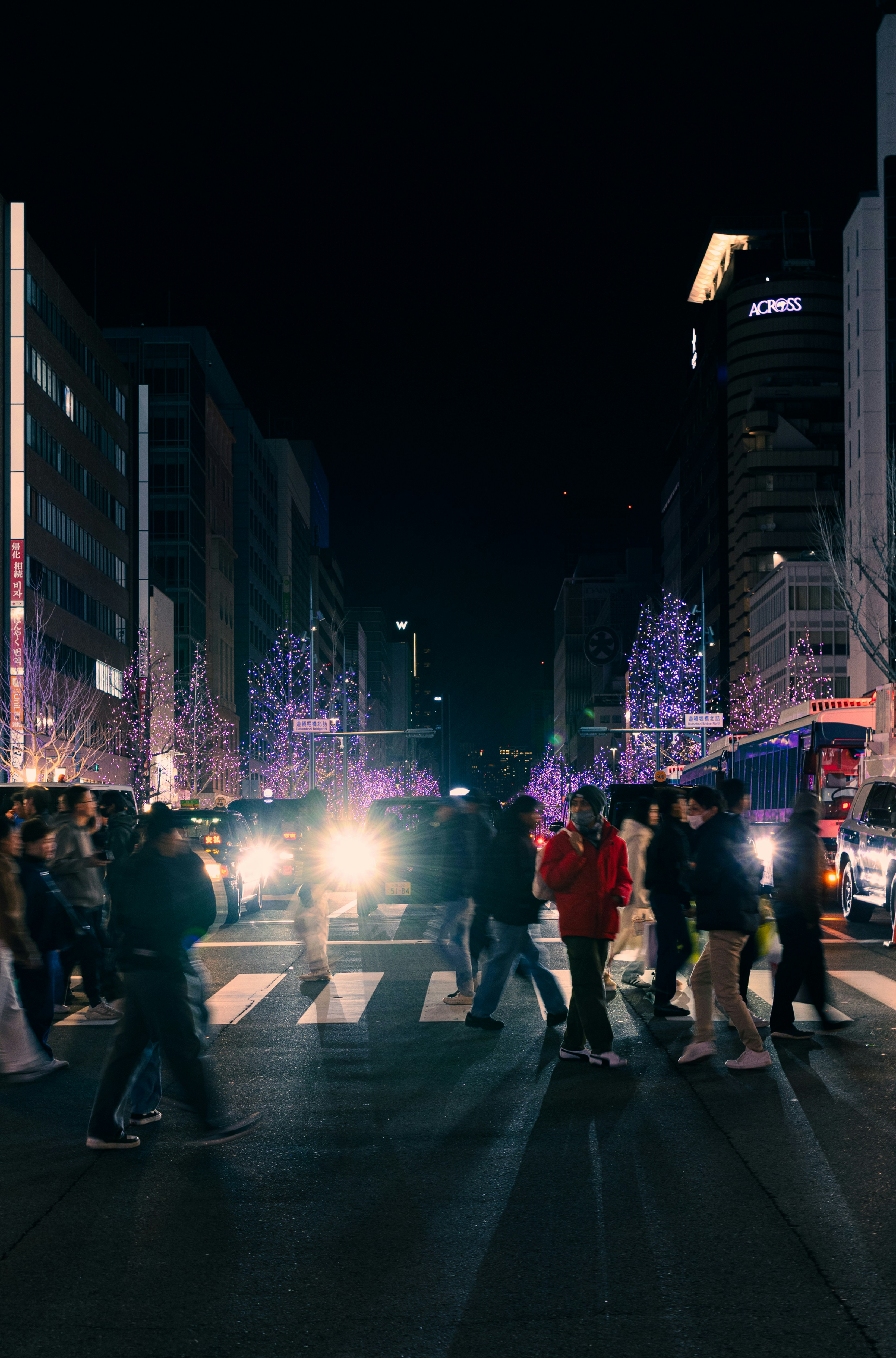 People crossing a street at night with lights.
