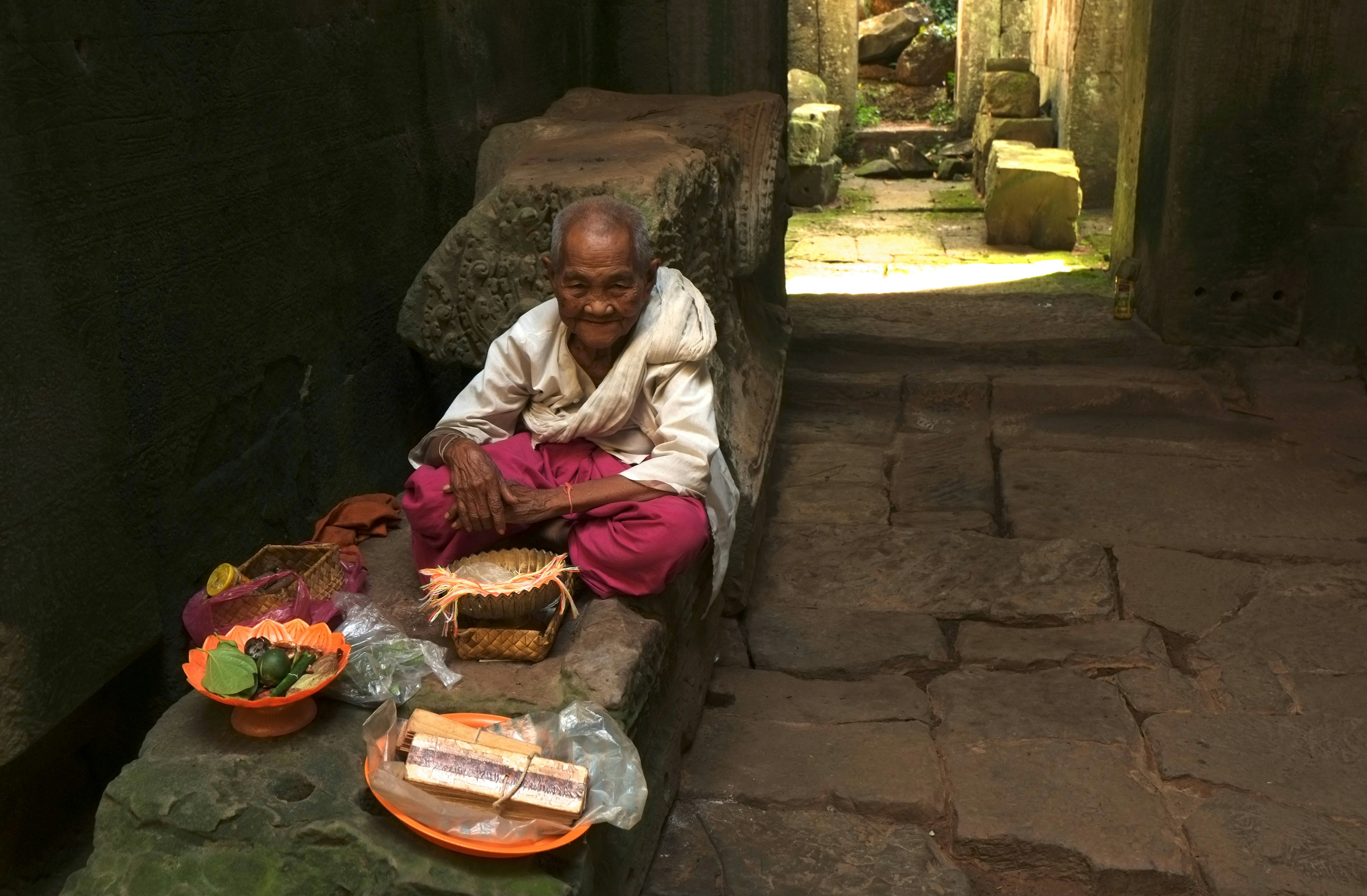 Elderly person sitting with offerings in ancient ruins.