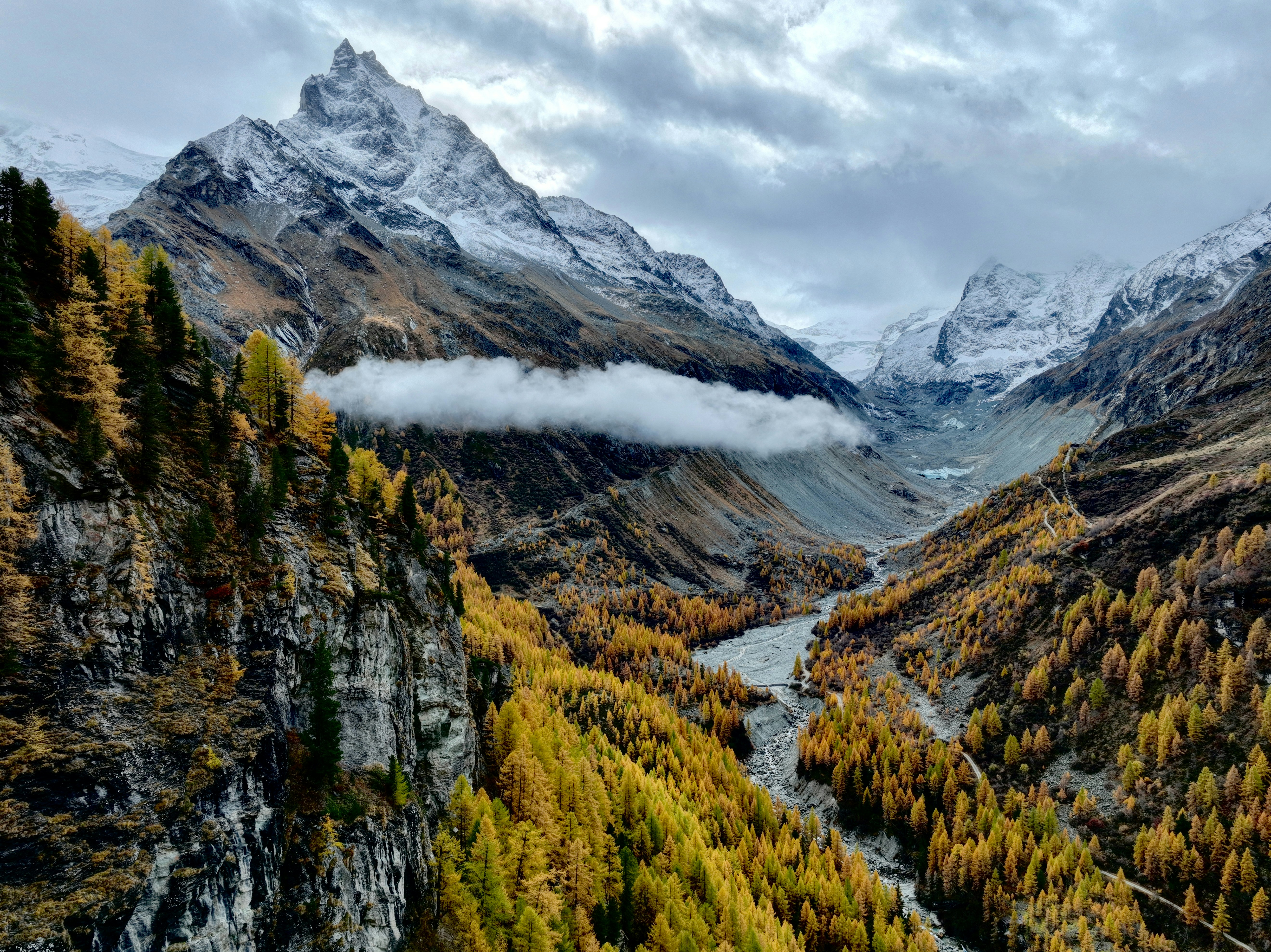Autumn trees line a mountain valley under snowy peaks.