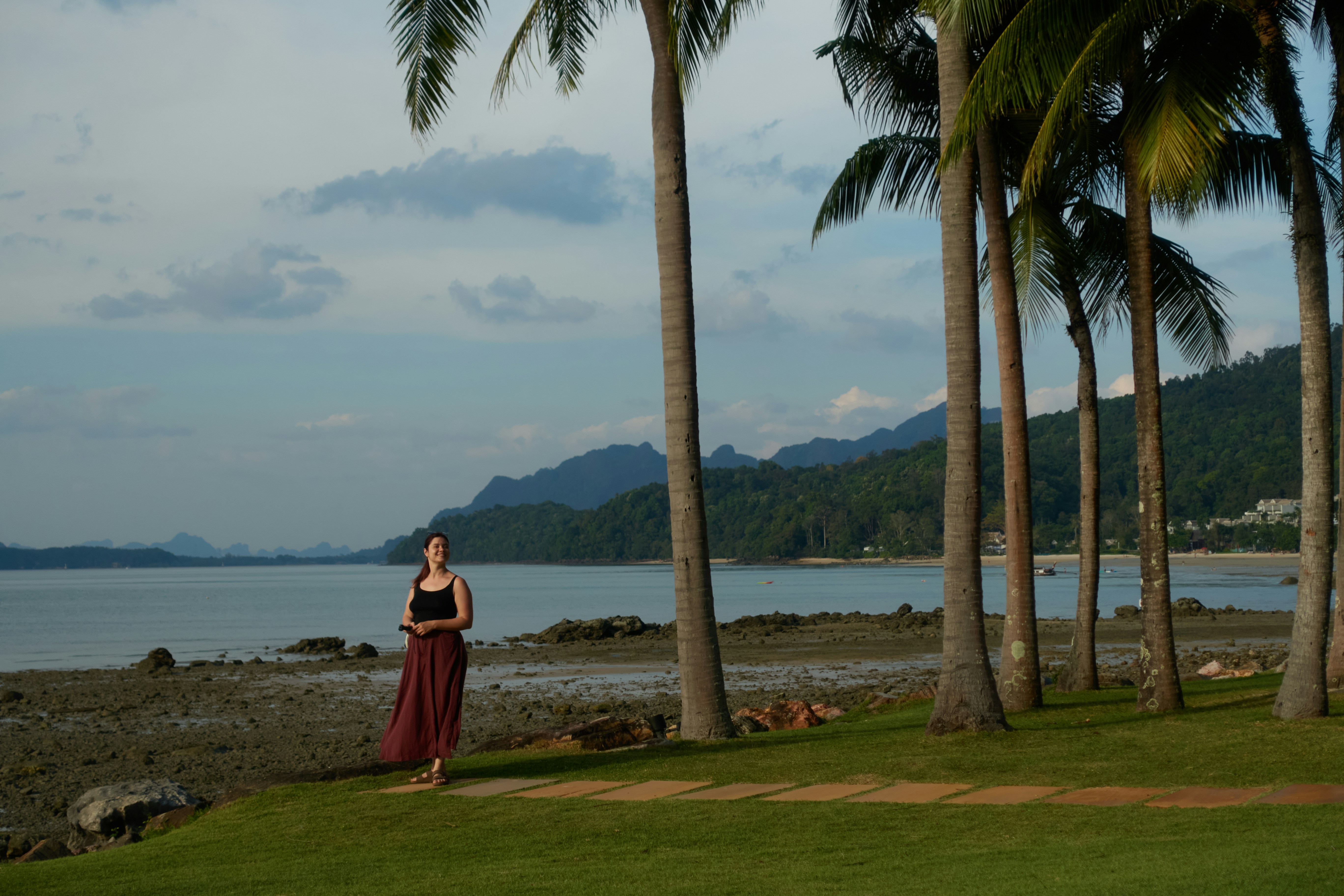 Woman standing on grass near palm trees by the ocean.