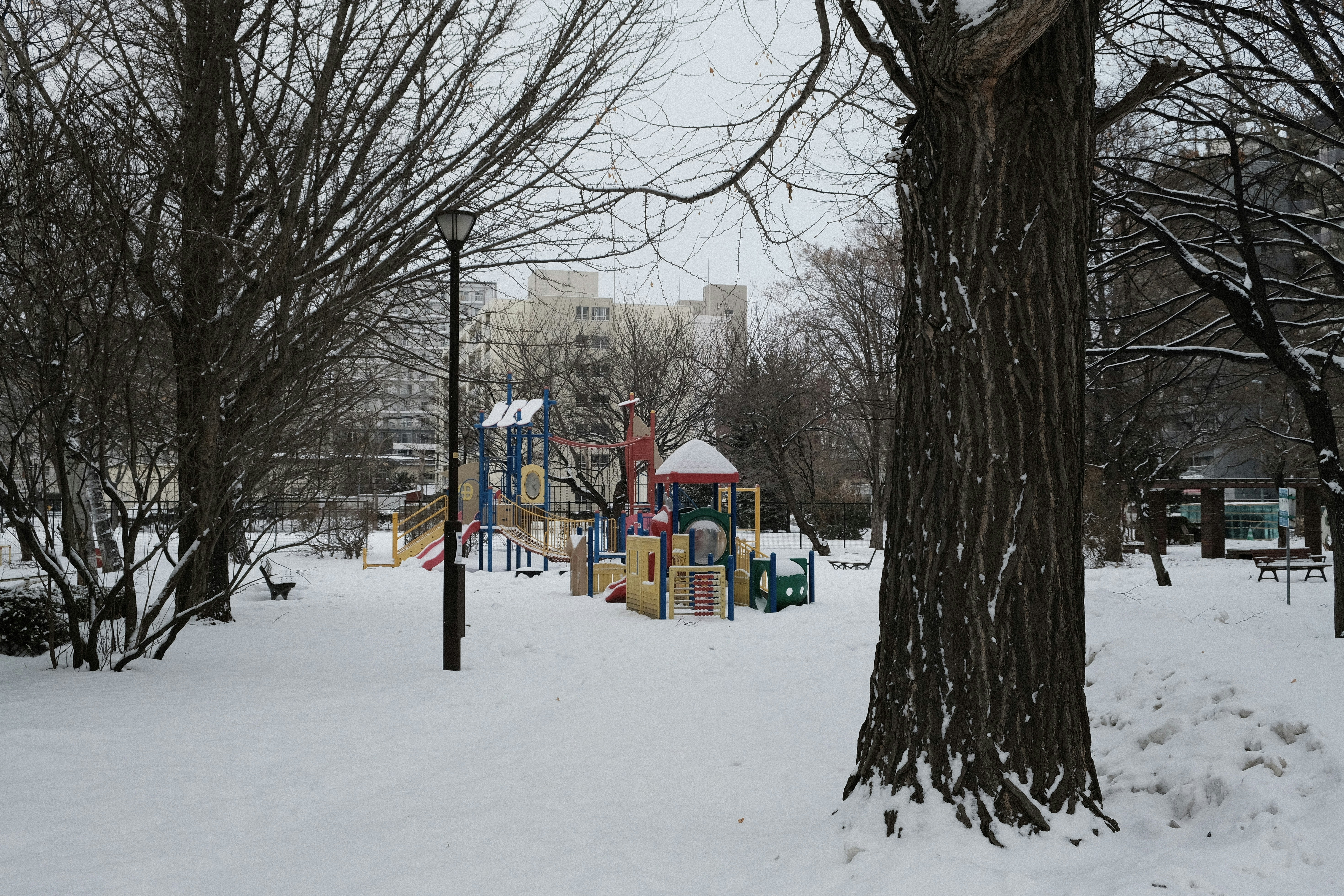 Snow-covered playground with bare trees and buildings.