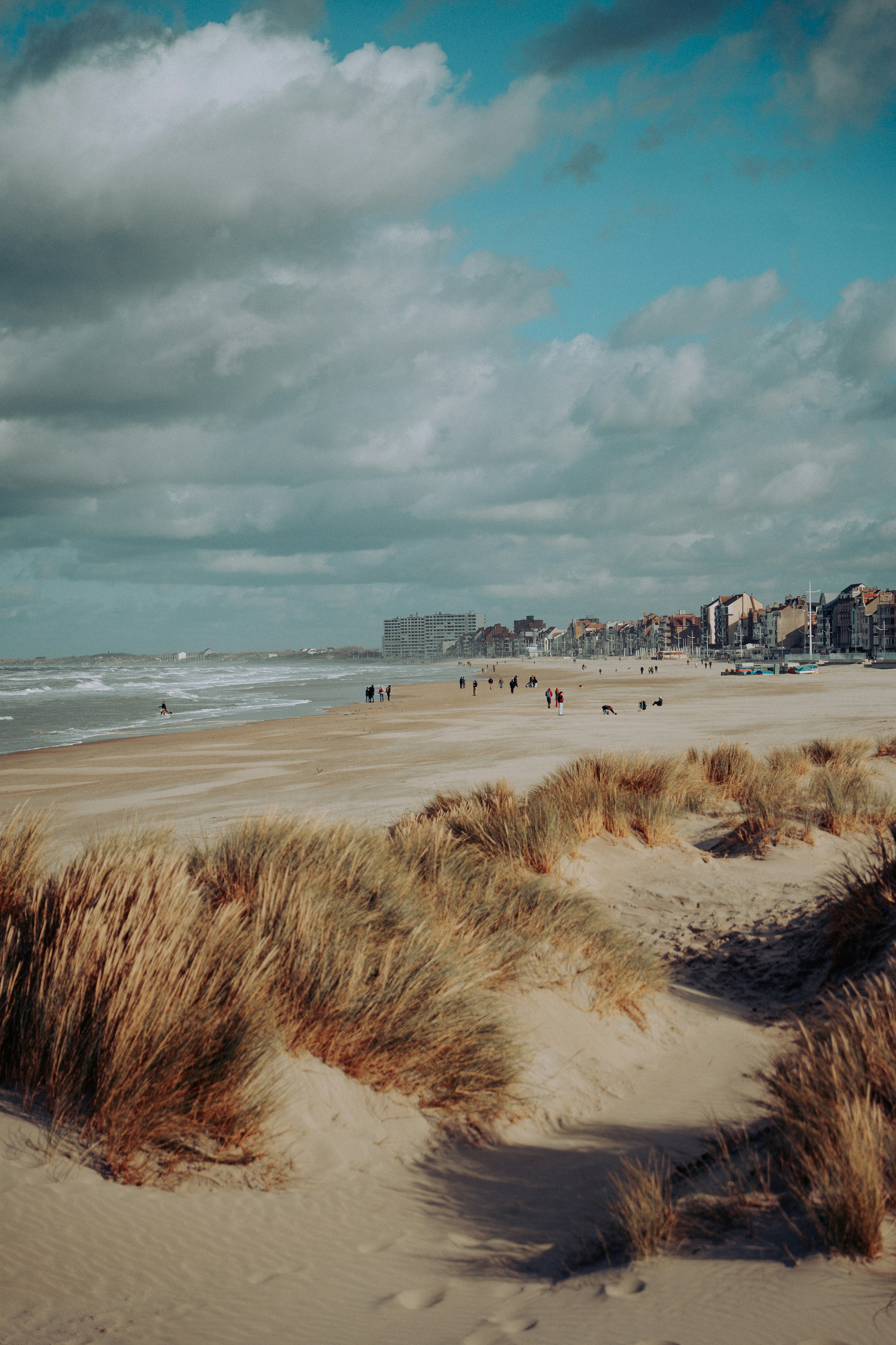 Dunas de areia e praia com prédios distantes sob céu nublado.