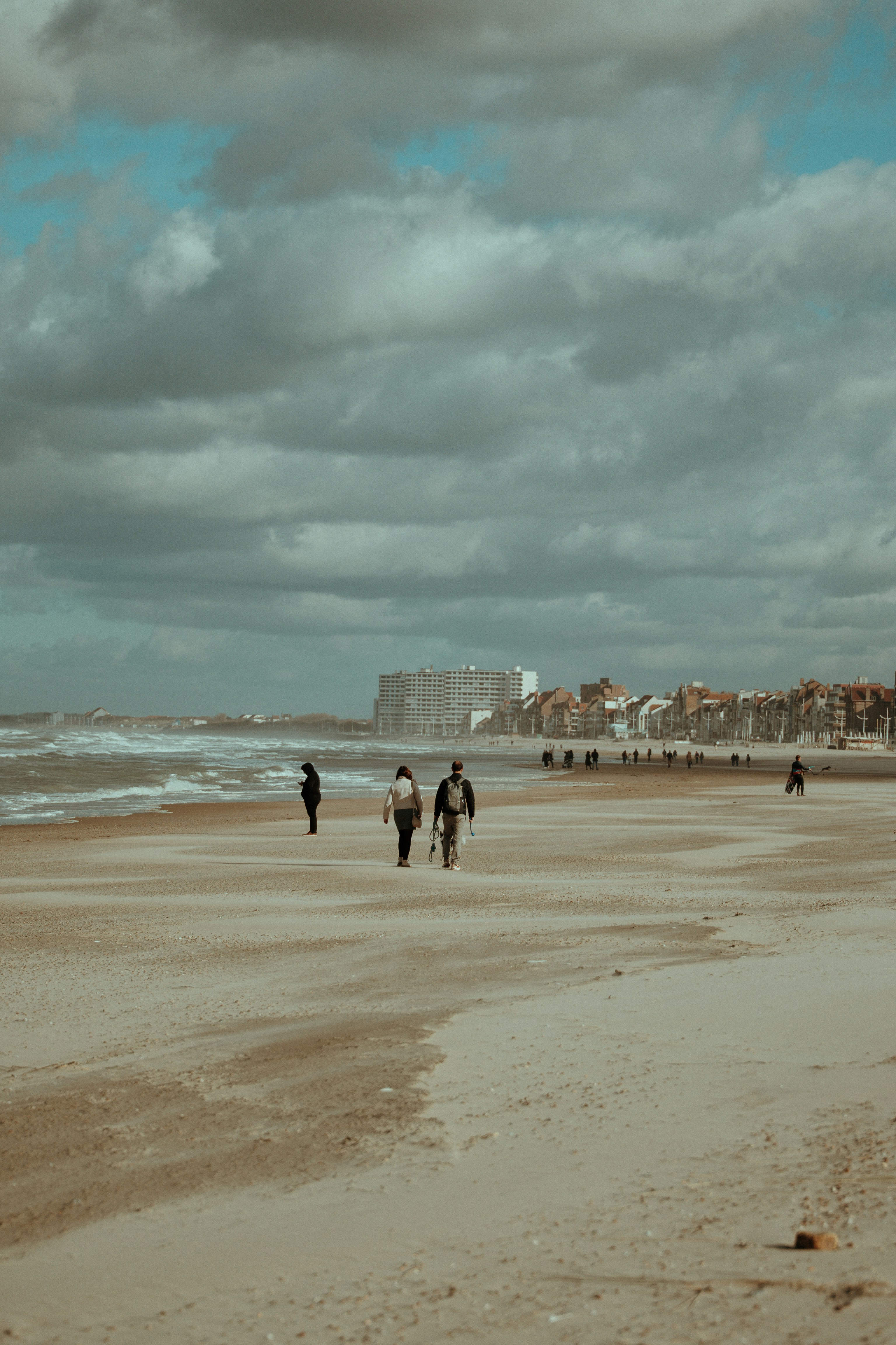Pessoas caminhando em uma praia de areia com prédios ao fundo.
