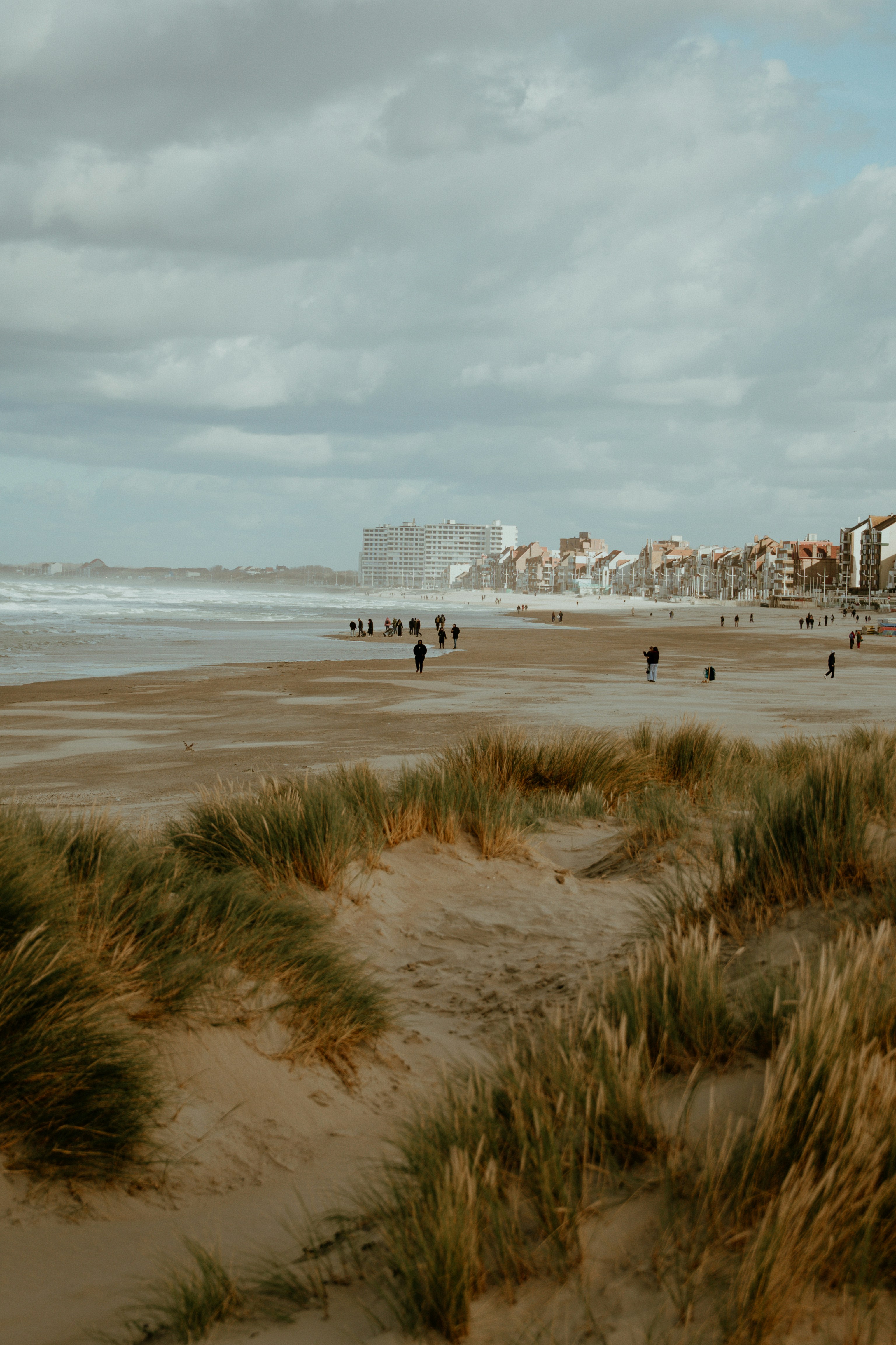 As pessoas caminham por uma praia de areia com dunas e prédios.