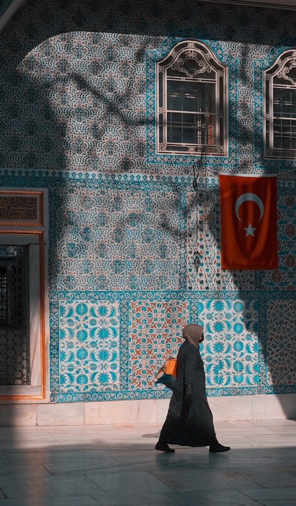 Woman in hijab walks past tiled wall with turkish flag.
