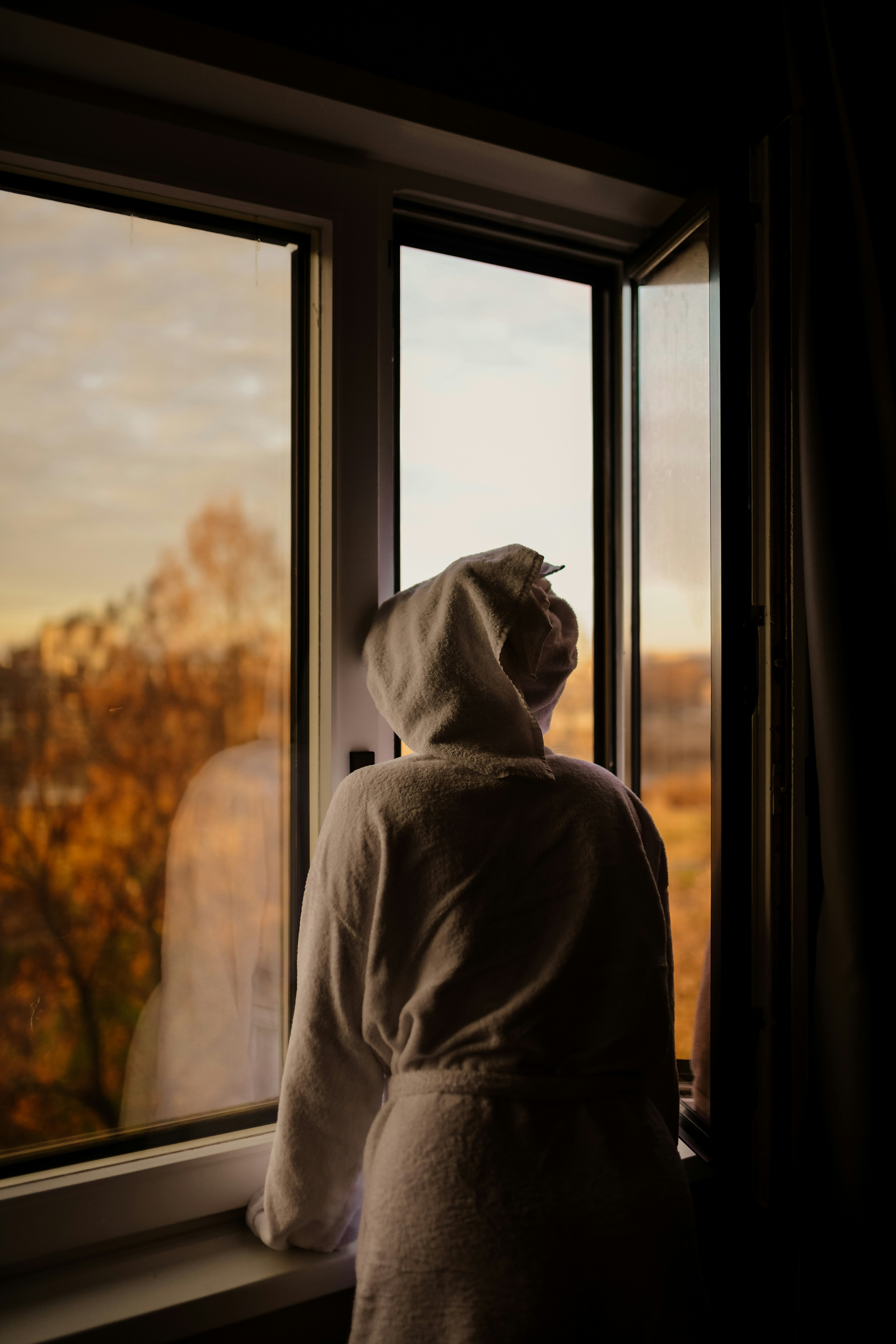 Person in bathrobe looking out window at autumn trees.