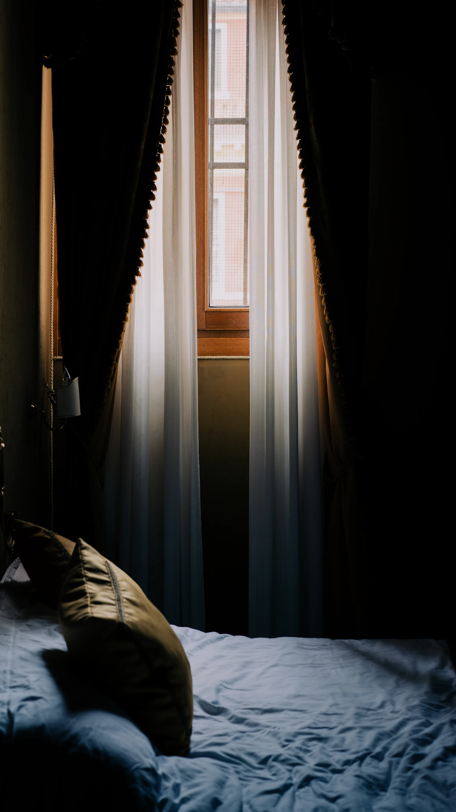 A dark bedroom with a heavy curtain pulled back from a bright window