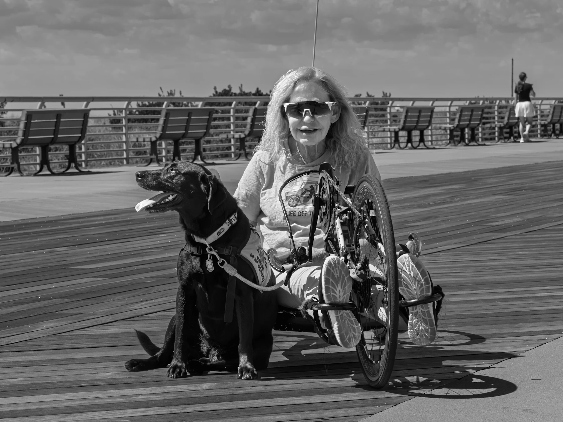 A woman on a handcycle beside her black service dog on a seaside boardwalk, in black and white