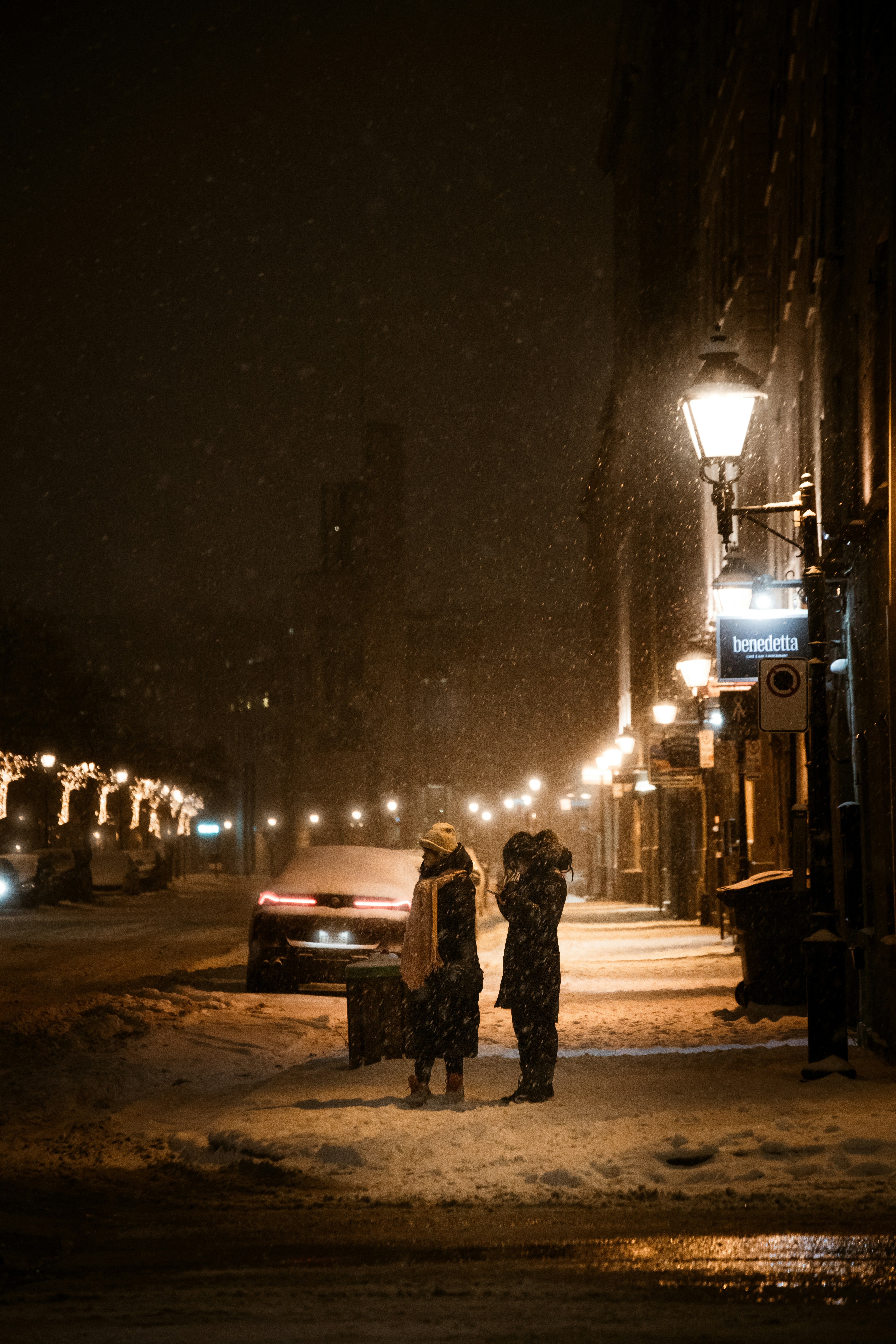 Two people stand on a snowy street at night.