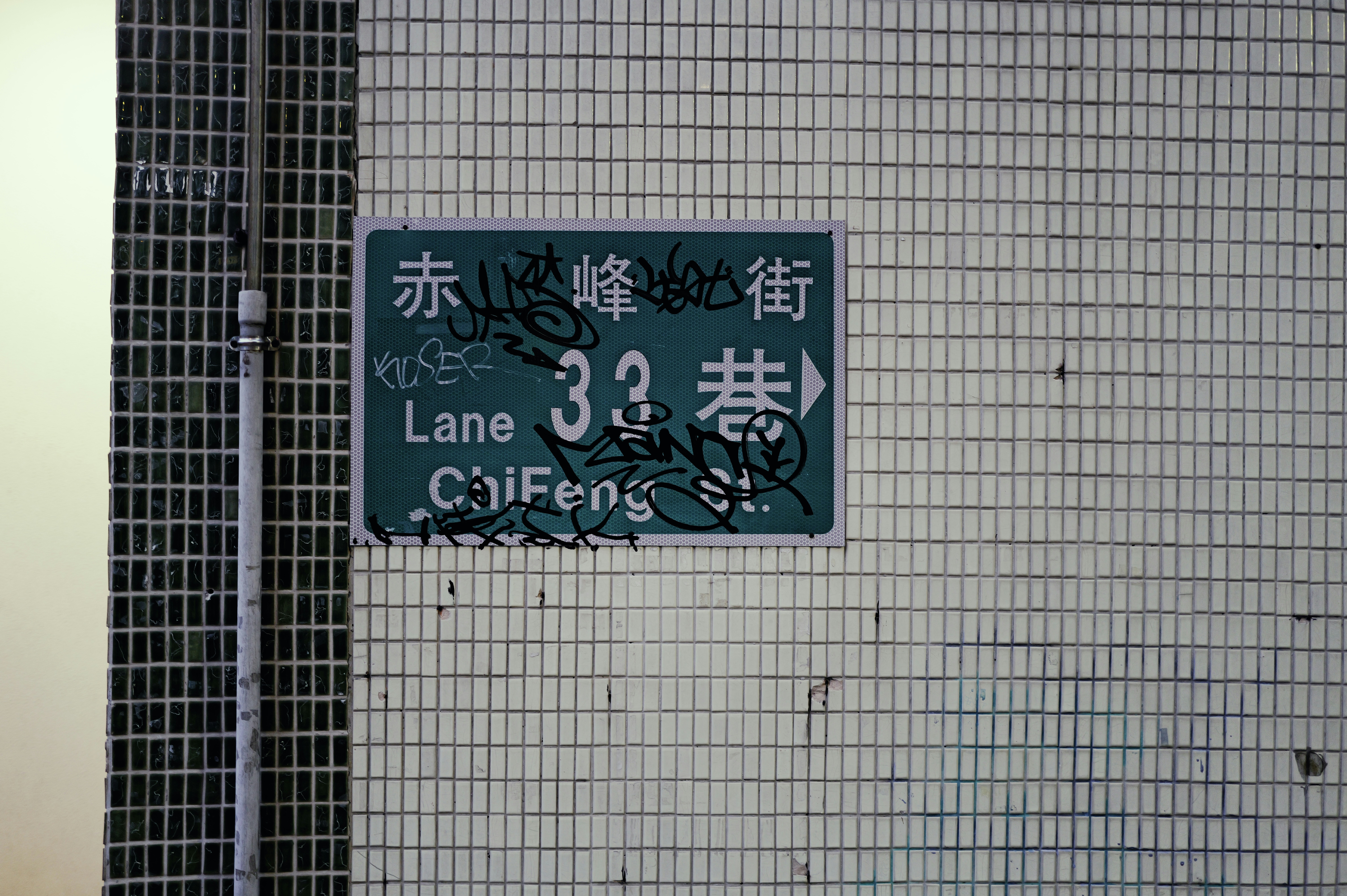 Green street sign with graffiti on tiled wall.