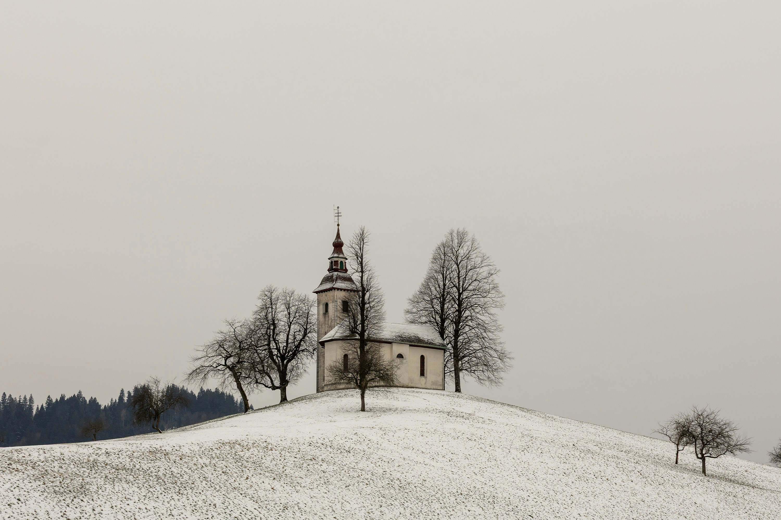 Eine kleine Kirche auf einem verschneiten Hügel mit kahlen Bäumen.