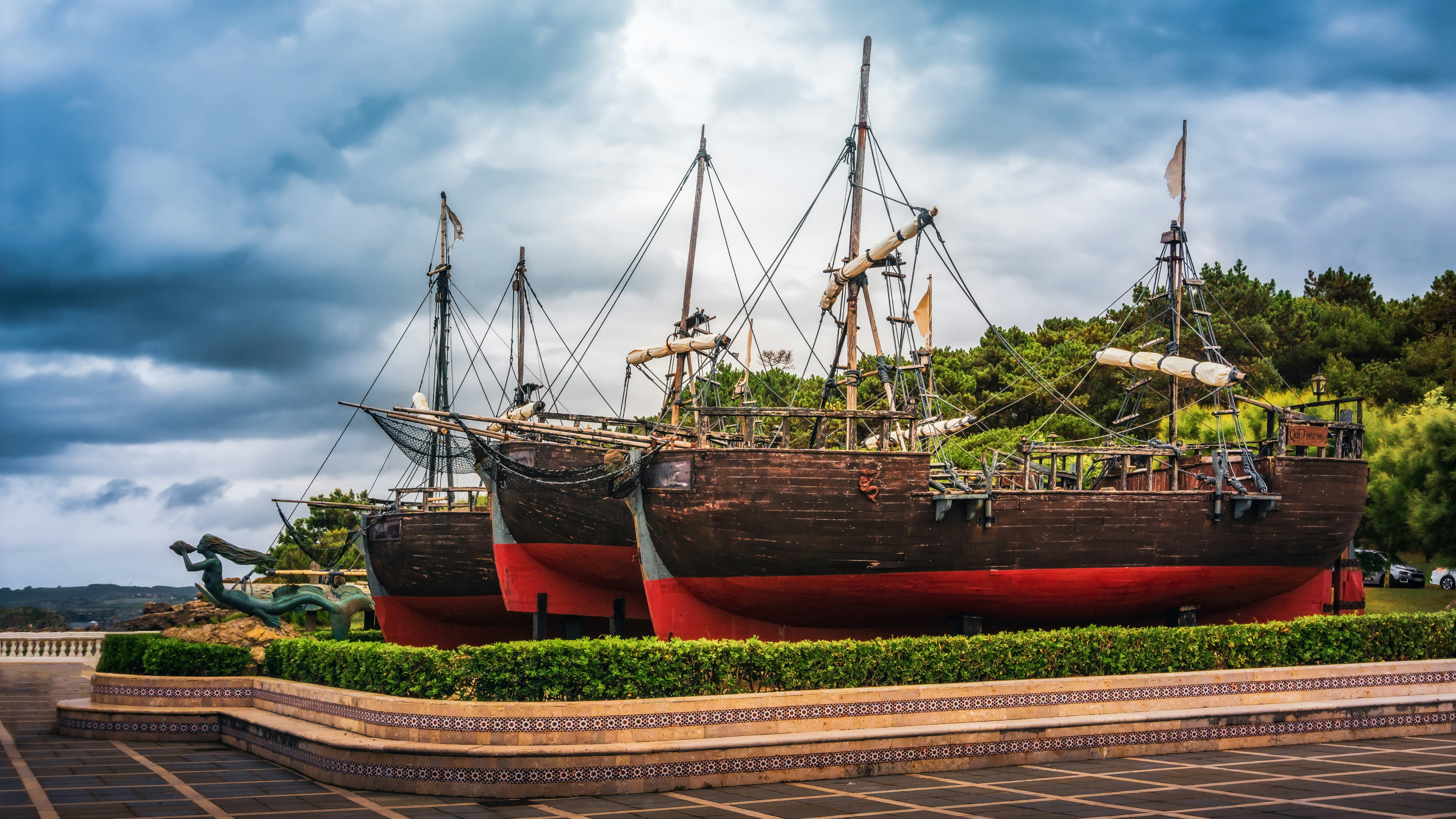 Three antique wooden ships docked near lush green trees.