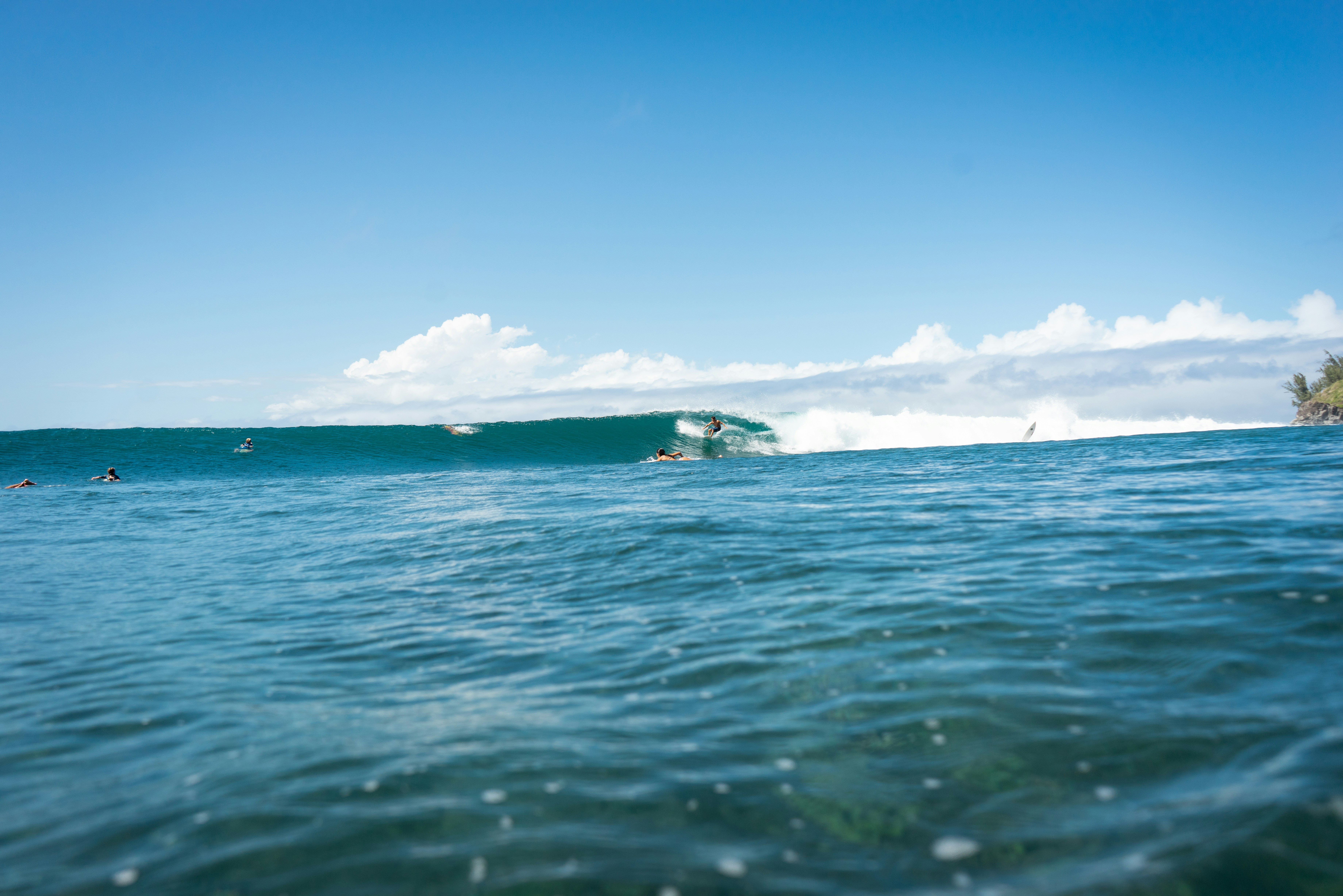 Surfers ride a large wave under a clear blue sky.
