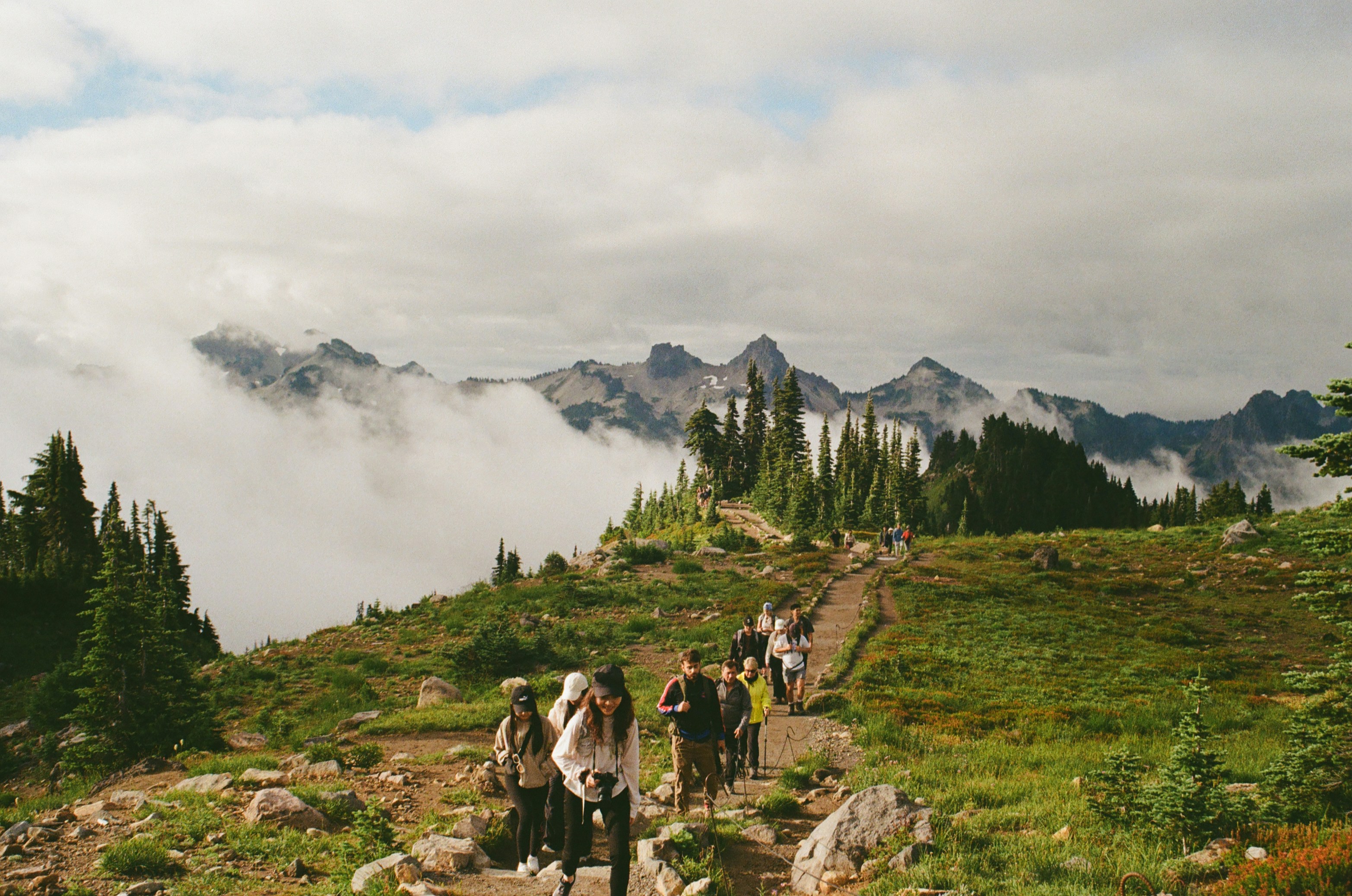 Hikers on a mountain trail with cloudy peaks.