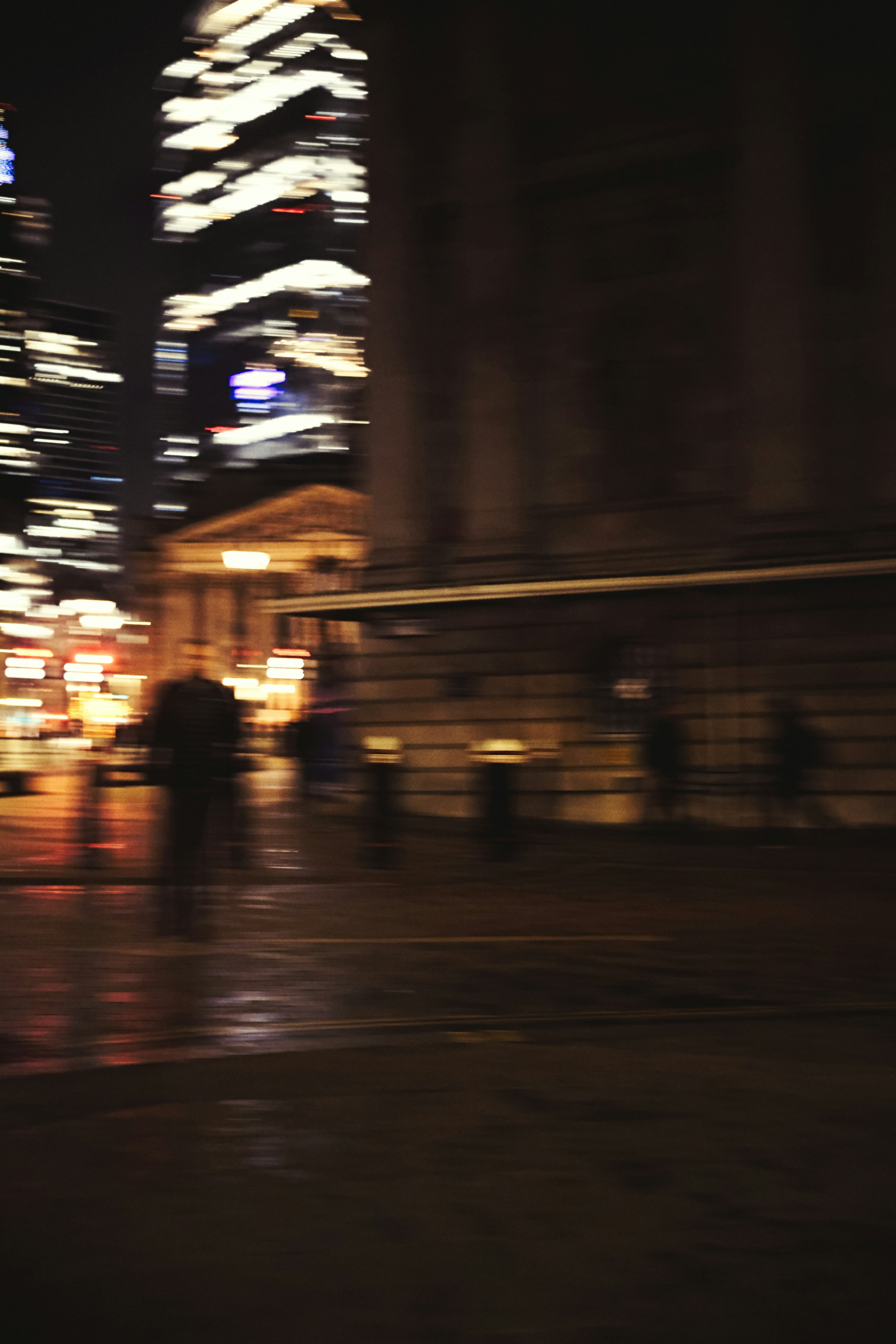 Blurred figures walk on a wet city street at night.
