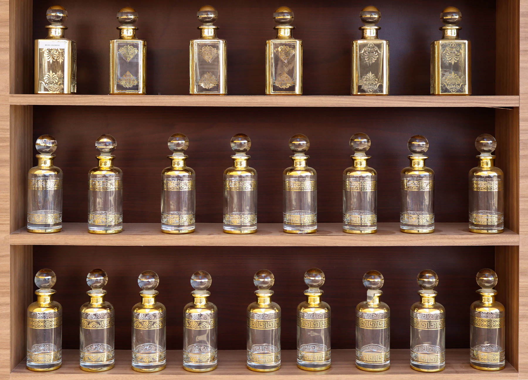 Three wooden shelves of matching clear glass bottles with gold collars and round stoppers, standing upright in even rows