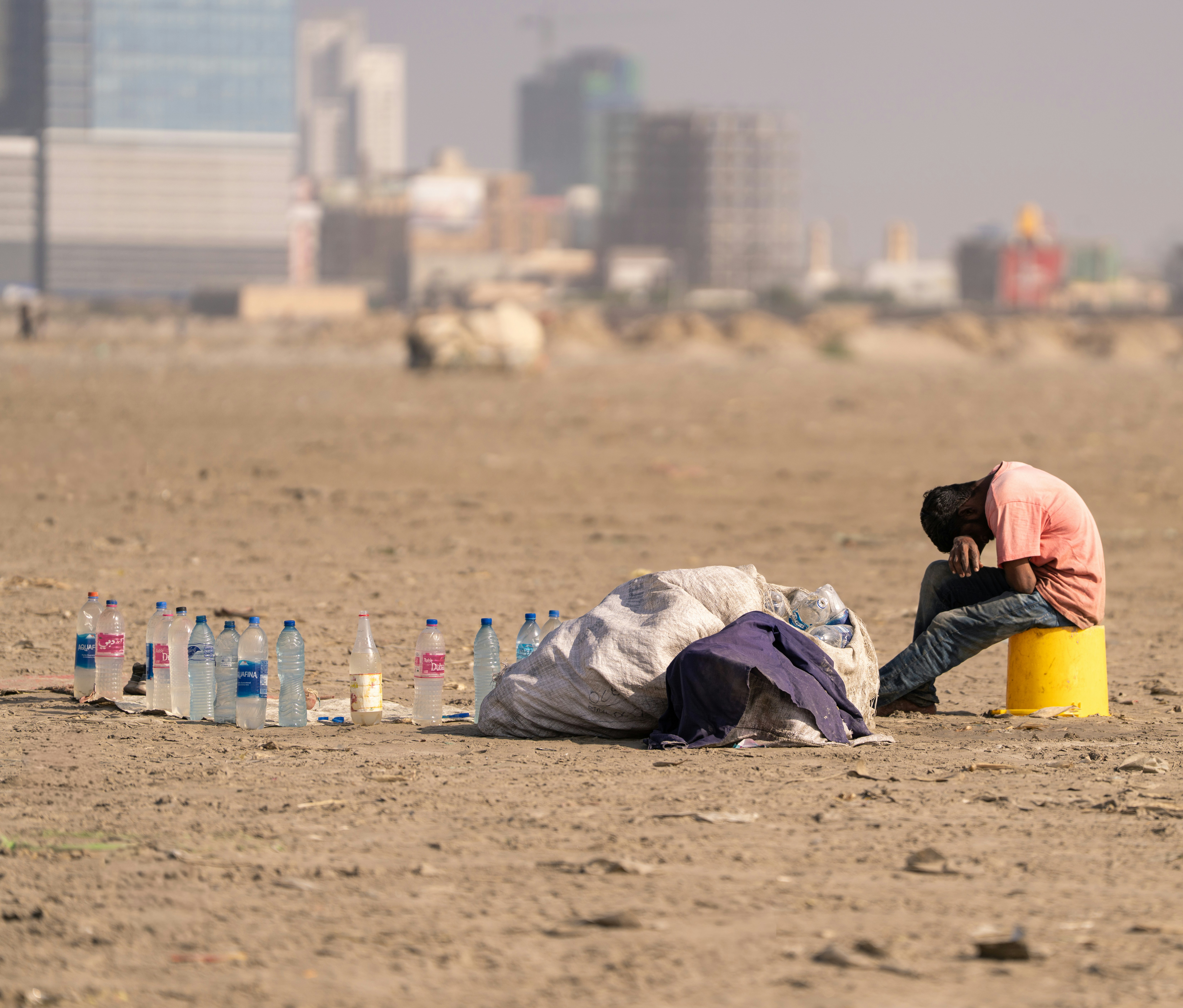 Street Worker Rests Beside Collected Plastic Bottles on a Beach with City in Background An exhausted street worker rests while sitting on a plastic container beside collected plastic bottles on a sandy beach, with a modern city skyline visible in the background. The image reflects urban struggle, informal labor, recycling work, poverty, and social inequality in a developing city environment. Captured in natural light with shallow depth of field.