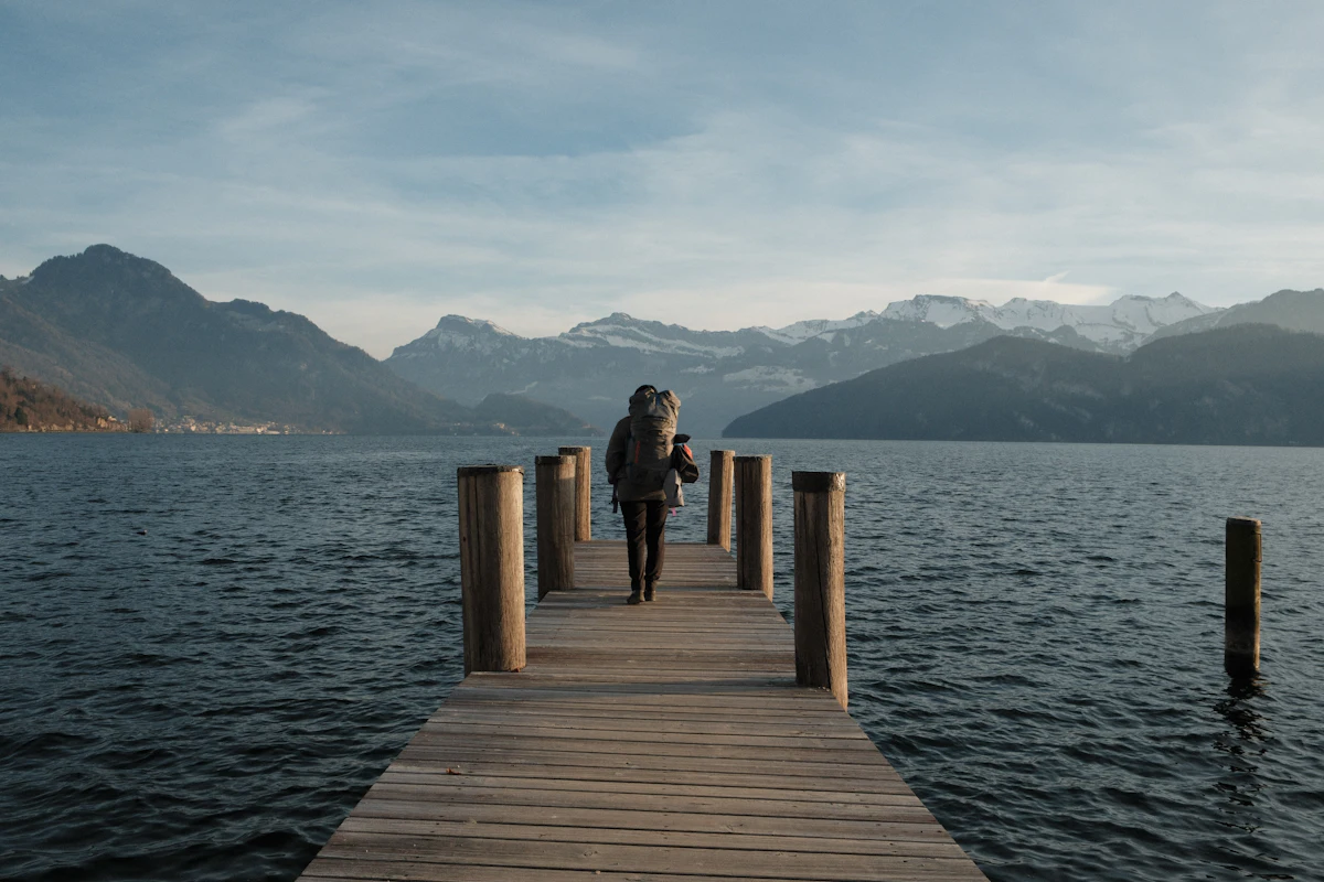 Solo traveler with a backpack walking down a wooden pier over a lake toward snow-capped mountains