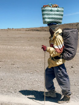 Man carrying basket and backpack on a dirt path.