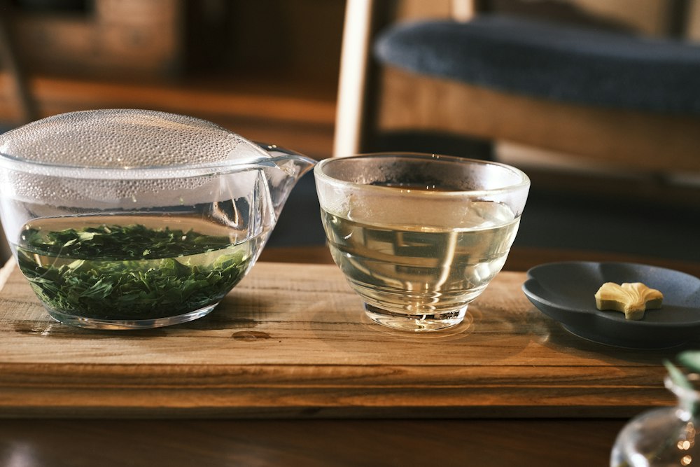 Clear green tea beside fresh sencha leaves in a glass pot on a wooden counter