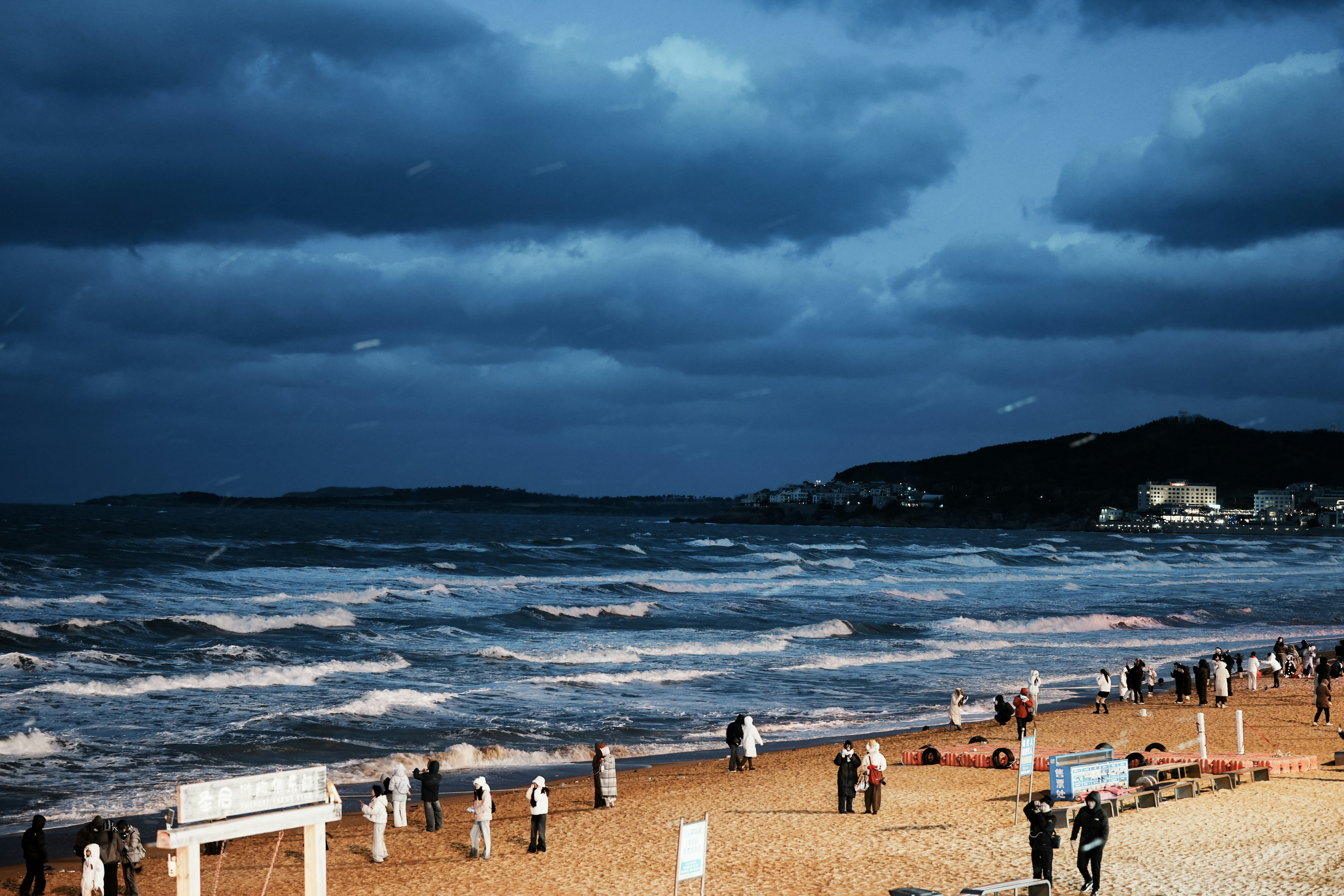 People walk on a sandy beach under dark clouds.